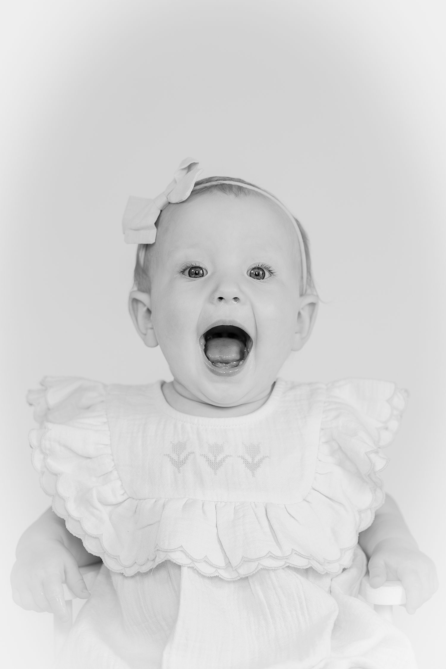 Black and white photo of a surprised baby girl in a white ruffled dress with a bow headband, mouth wide open.