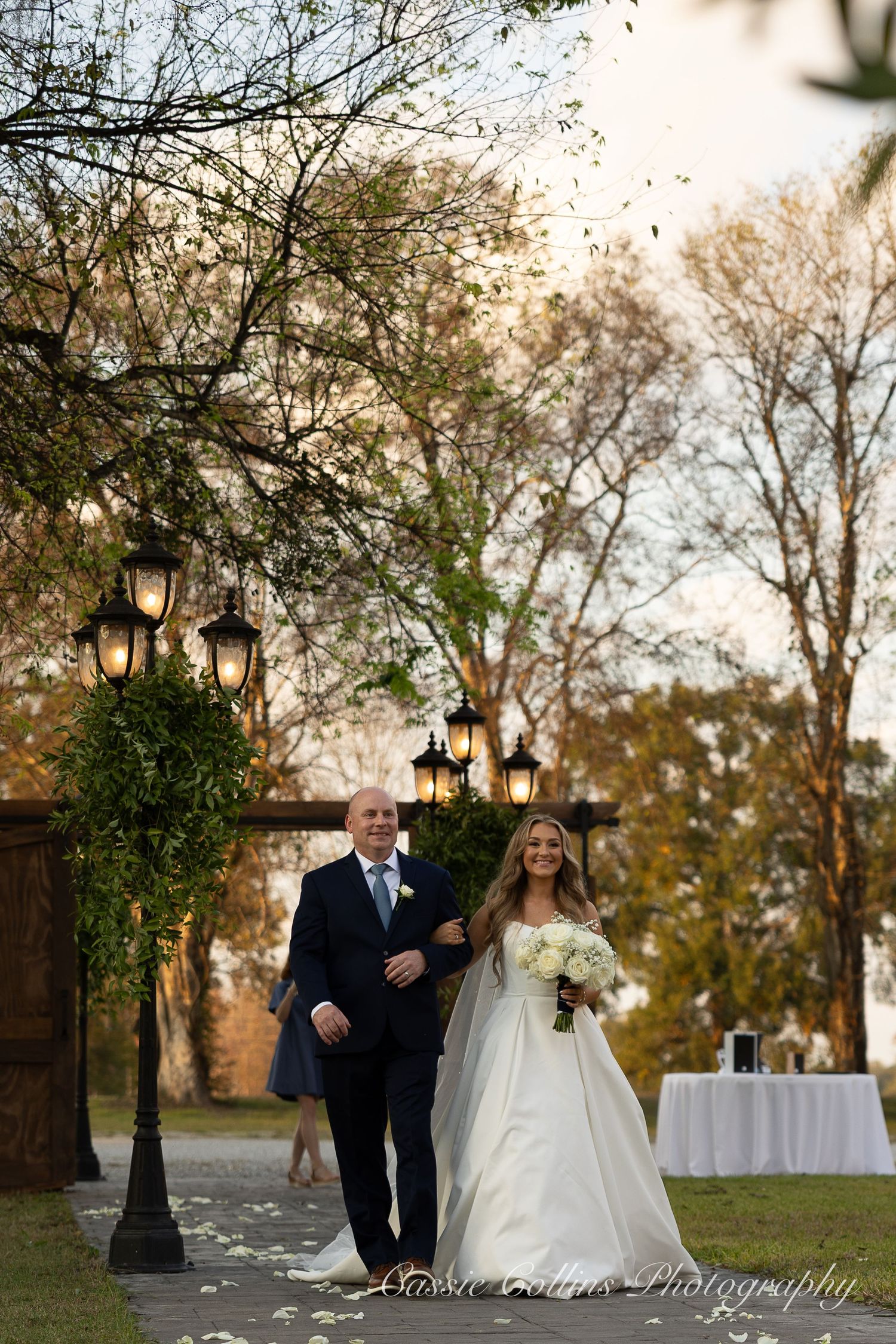 Romantic wedding couple walking along a path with lantern-lit trees at dusk creating a magical outdoor setting.