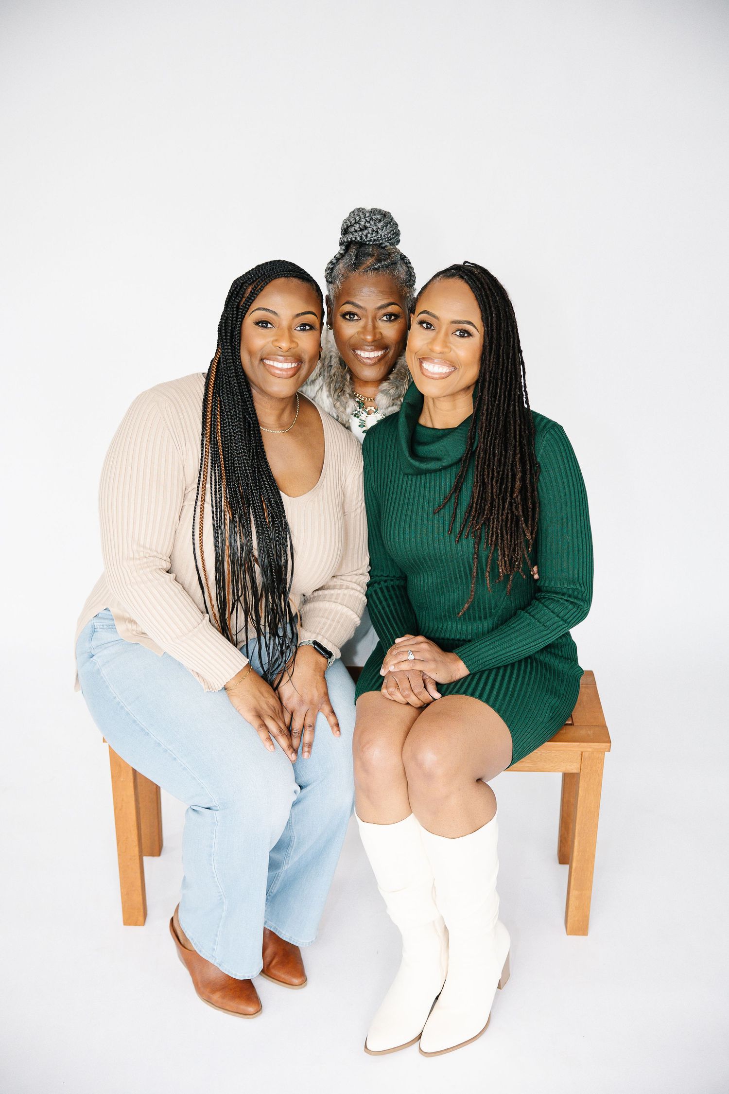 Three people sit together on wooden chairs against a white backdrop, wearing coordinated outfits.