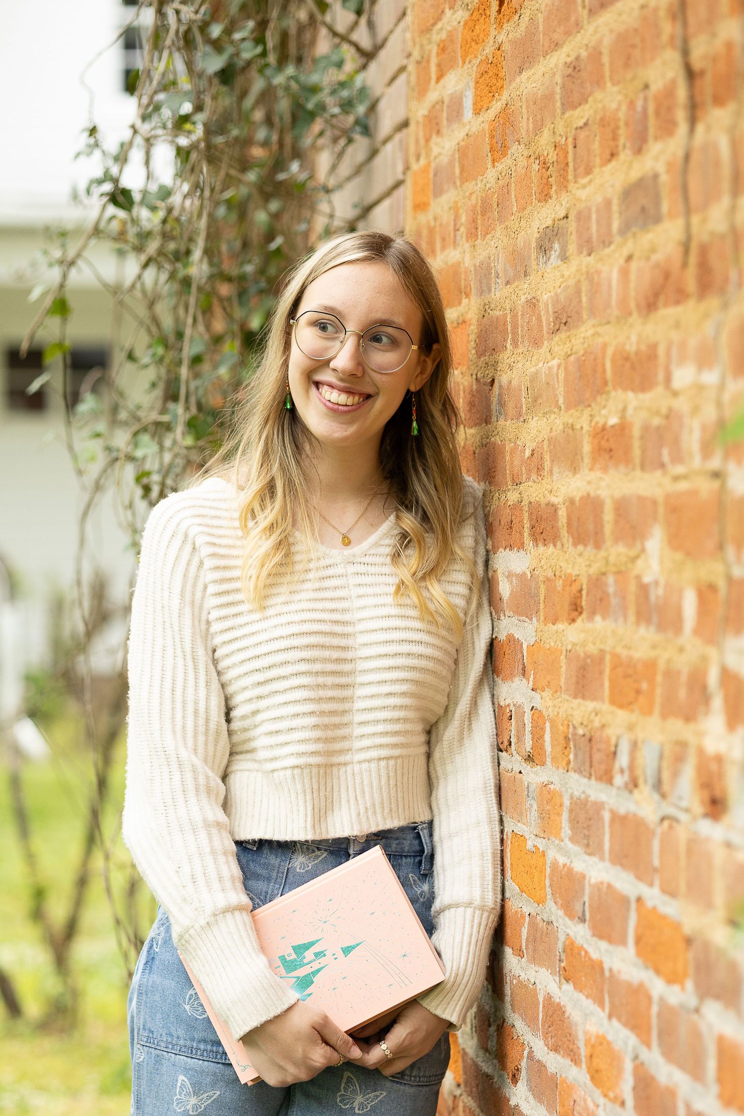 Smiling young woman with glasses holding a pink book, leaning against a brick wall in a cream knit sweater.