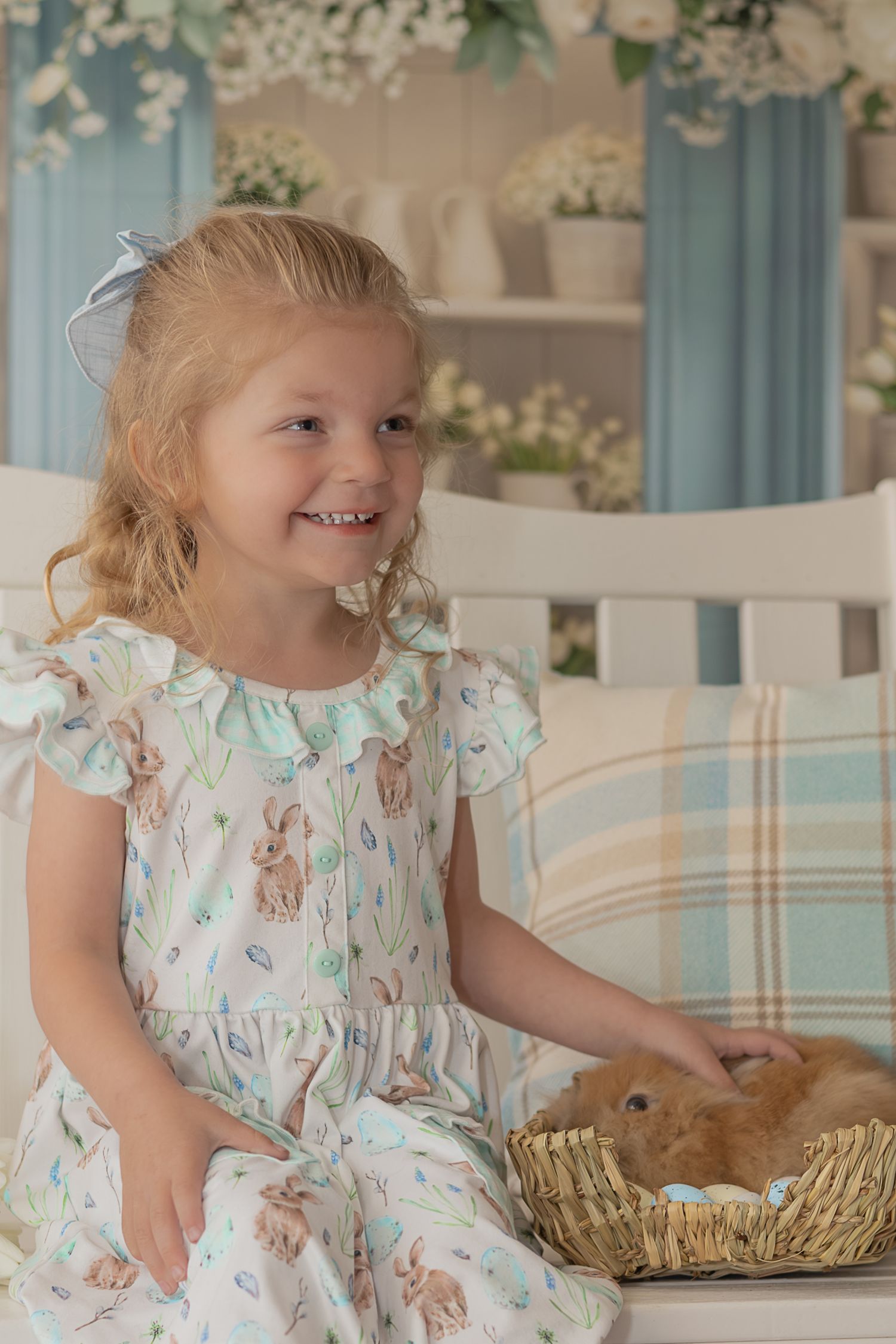 A child in a floral dress sits on a white bench next to a woven basket containing a small brown bunny rabbit.