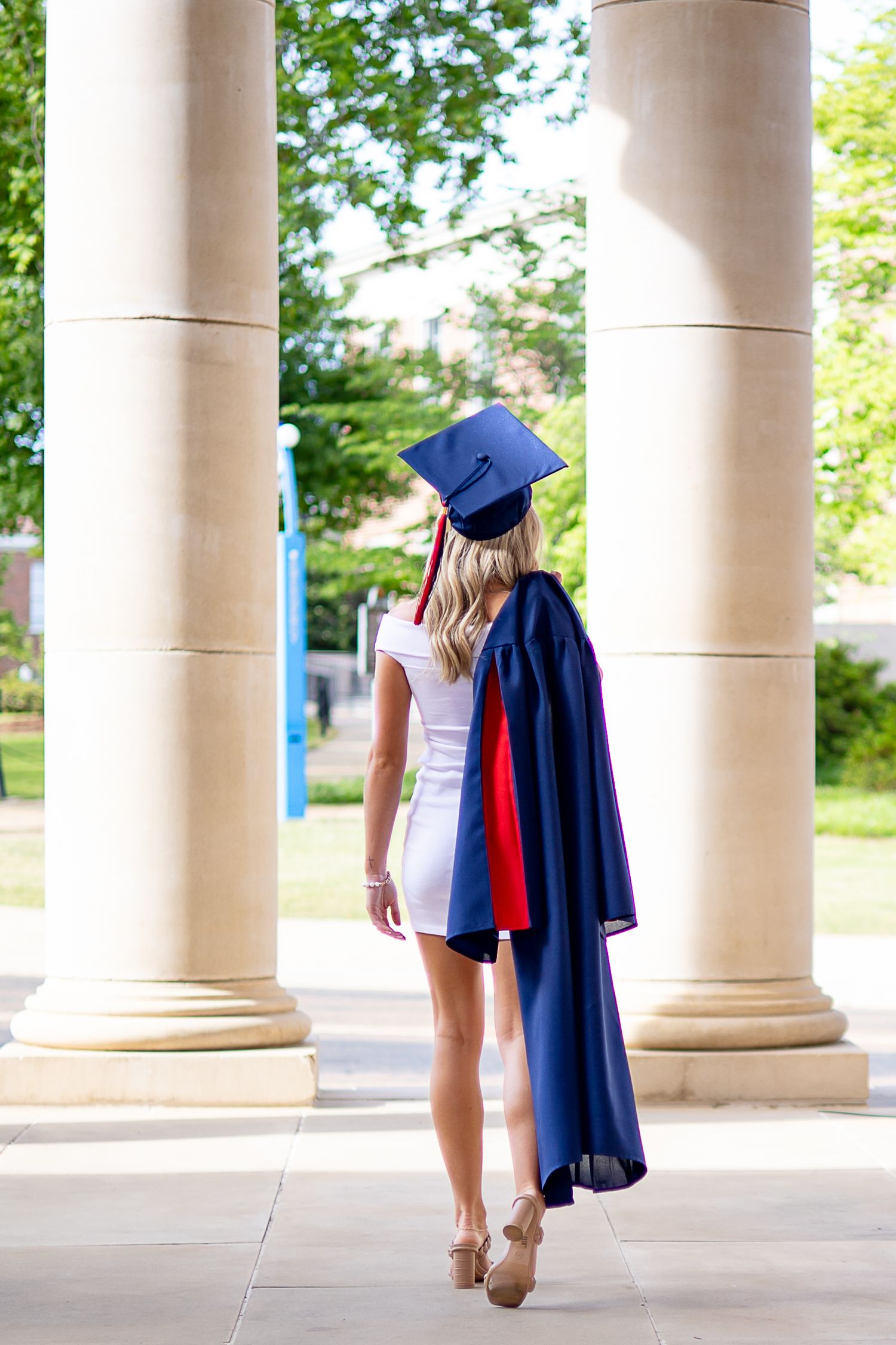 A female graduate in a blue cap and gown stands between stone columns, viewed from behind on a sunny campus.