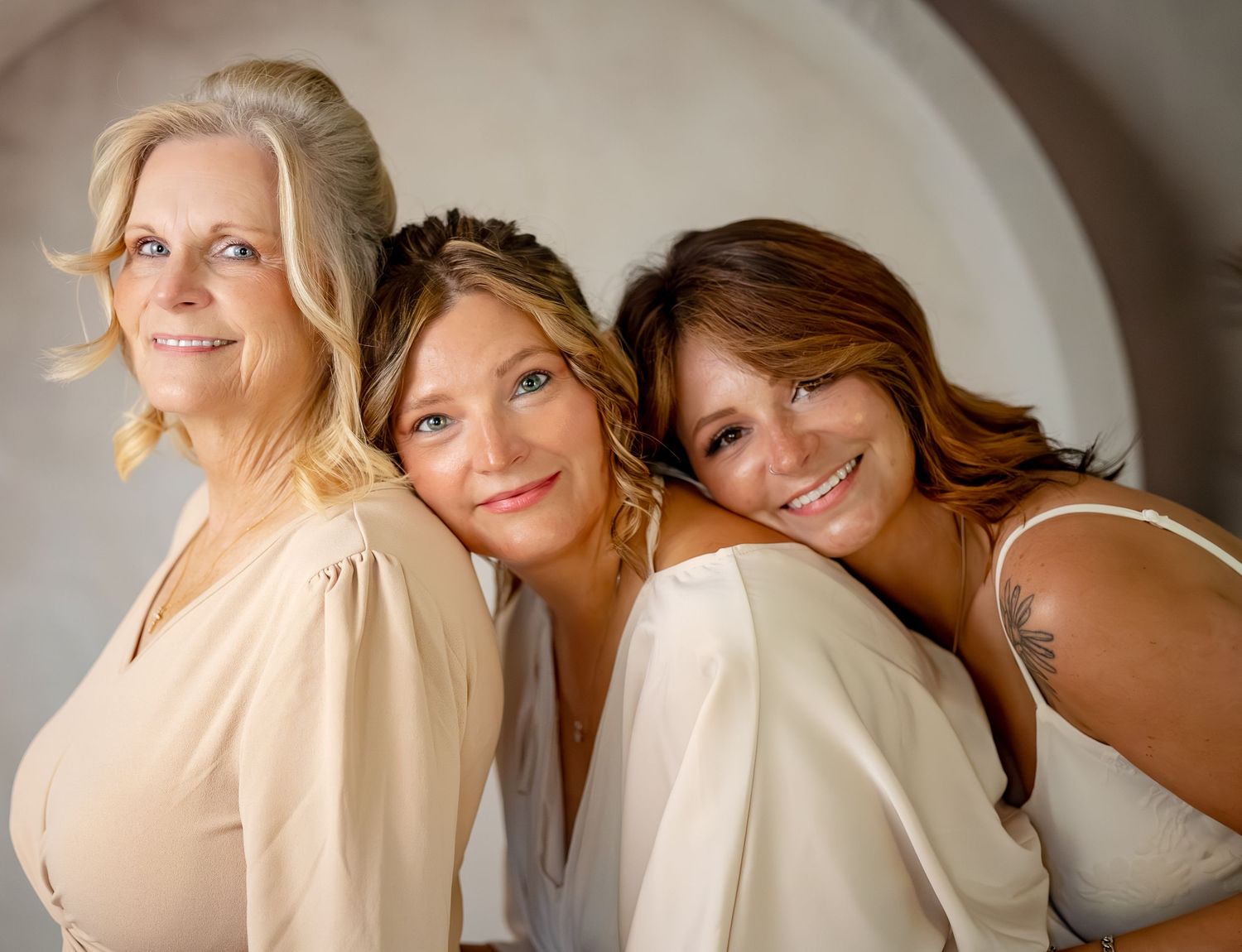 Three women of different ages pose together smiling, wearing neutral tones in a professional portrait photo.