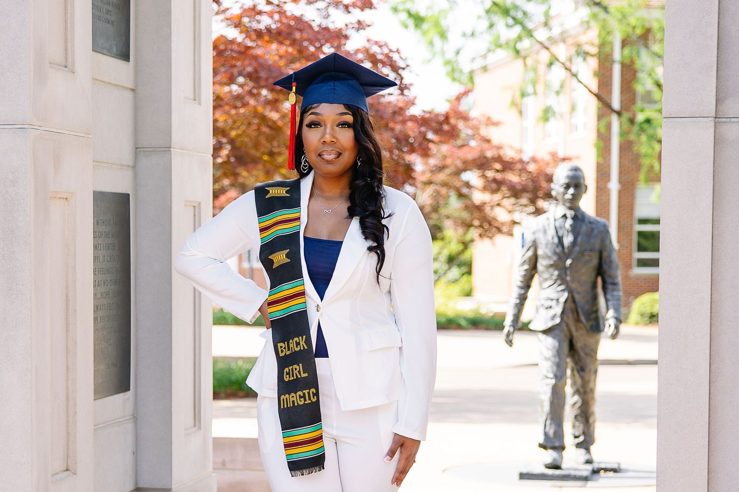 Graduate in white suit and cap with kente cloth stole poses proudly near a statue on a sunny campus.