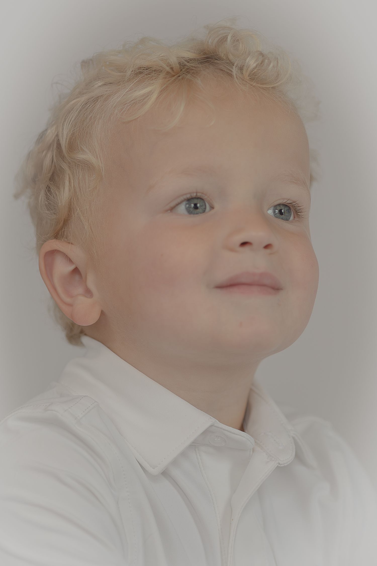 Smiling toddler boy with curly blonde hair and blue eyes wearing a white shirt in a soft portrait photo.