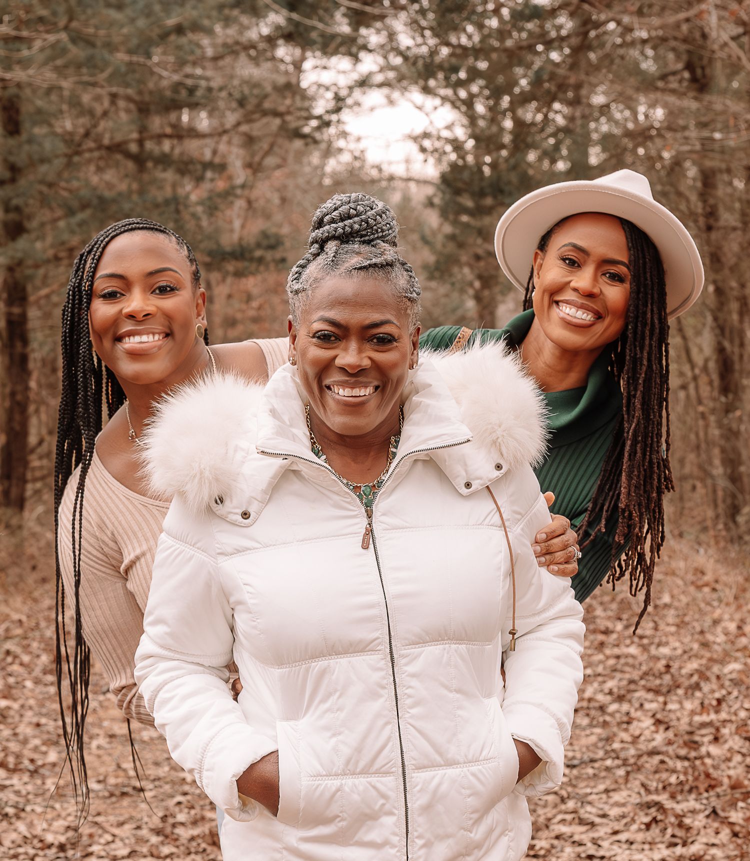 Three women in casual outdoor wear smile warmly while standing together in a wooded area during autumn.