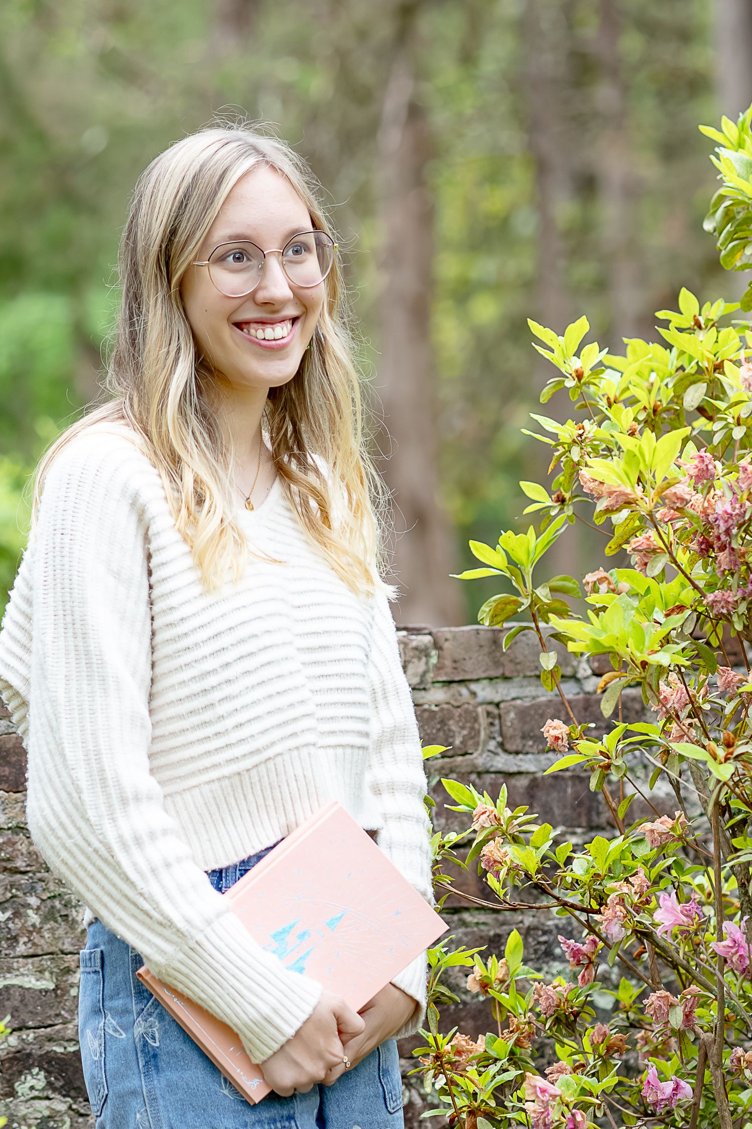 Smiling blonde woman with glasses holding a book outdoors near pink flowering shrubs in a white knit sweater.
