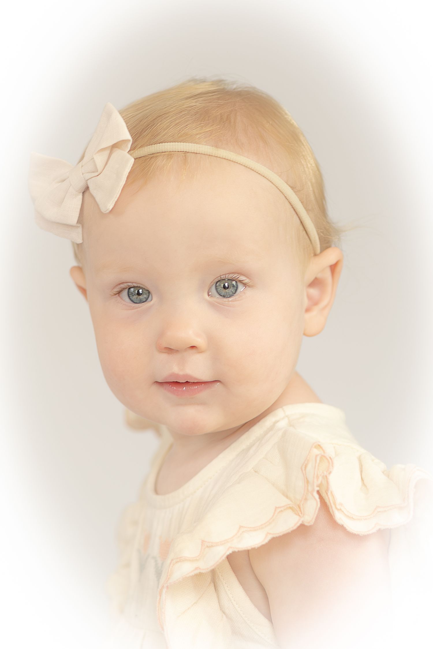 Adorable blonde baby girl with blue eyes wearing a cream dress and bow headband in a soft portrait photo.