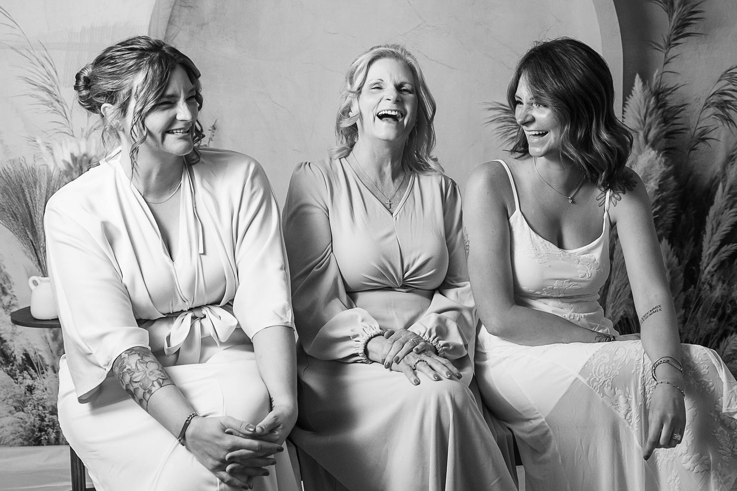 Three women in elegant attire laugh joyfully together in a black and white wedding photo.