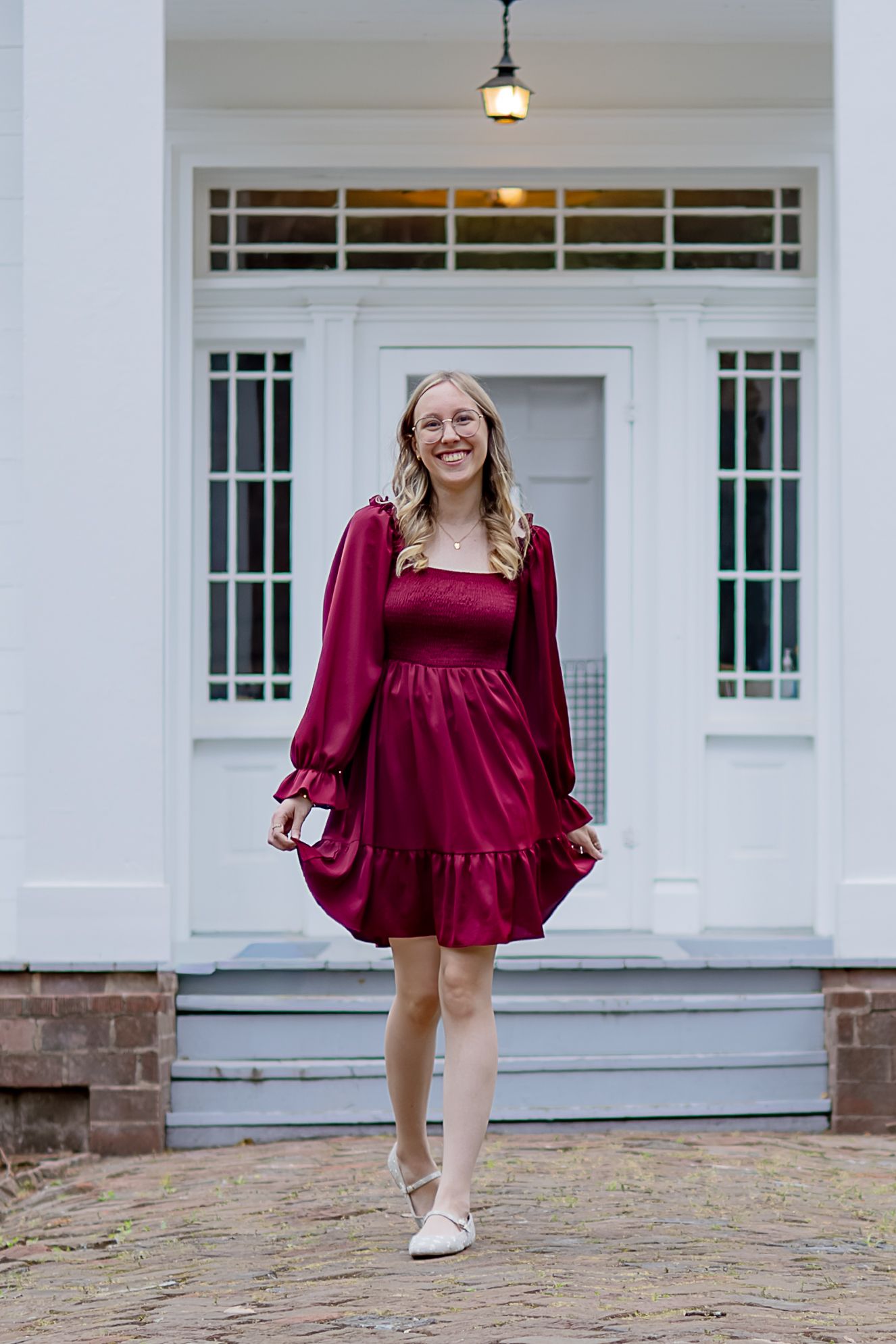 Young woman in a crimson satin mini dress with long sleeves smiling outdoors in front of a white building.