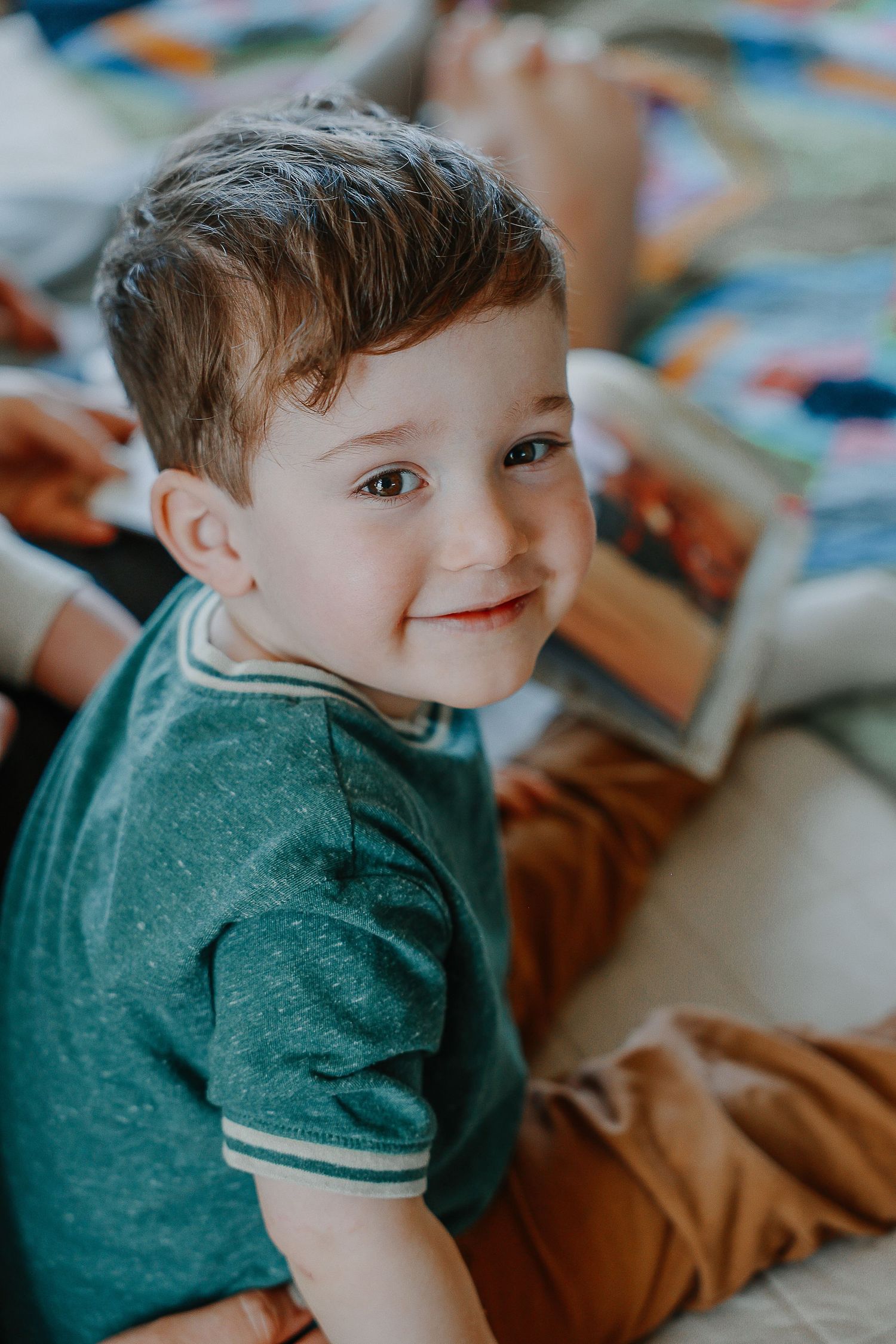 Adorable toddler boy in green shirt smiling warmly while sitting on a bed with a colorful book nearby.