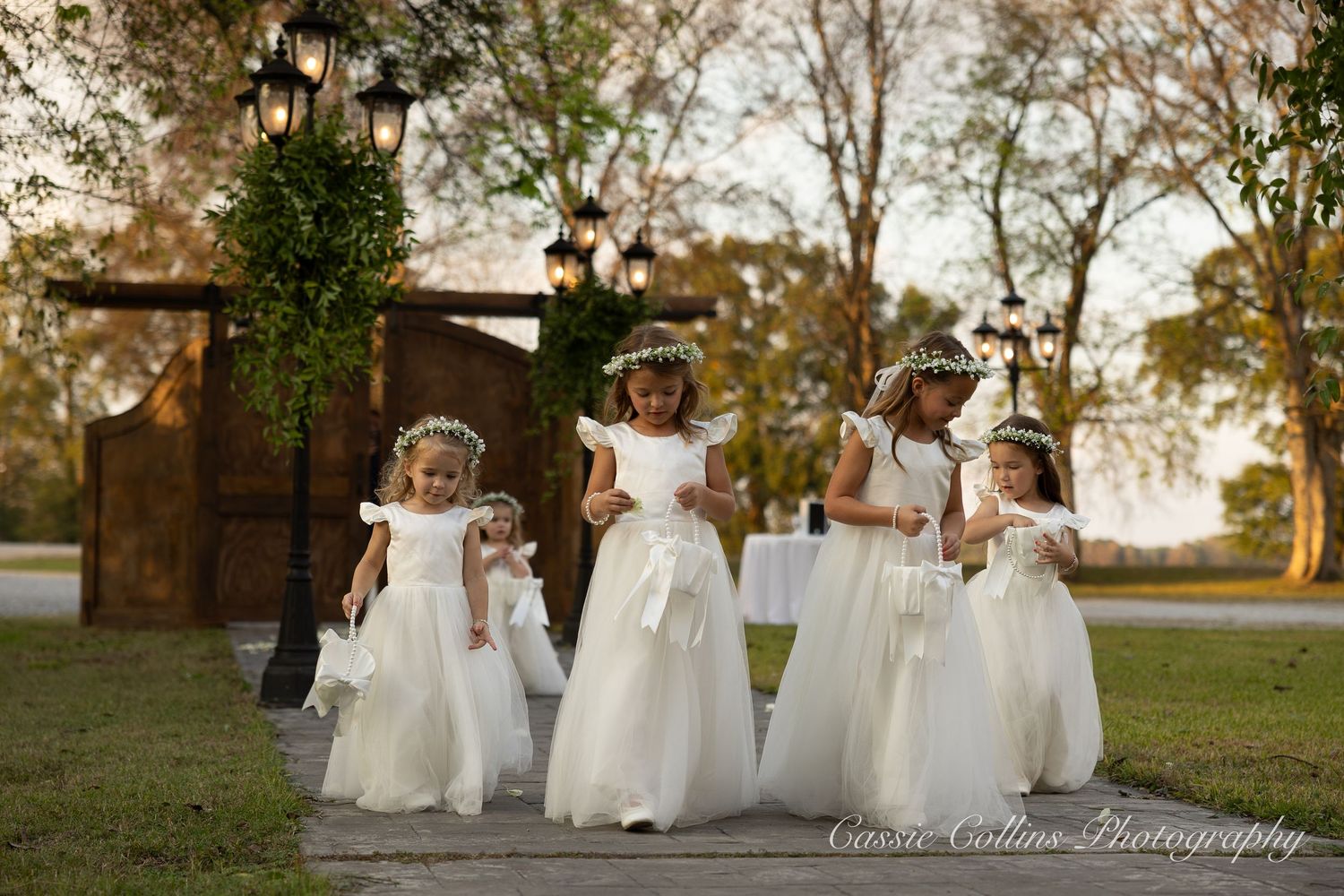 A group of people in white dresses walk together across a grassy outdoor setting at sunset.
