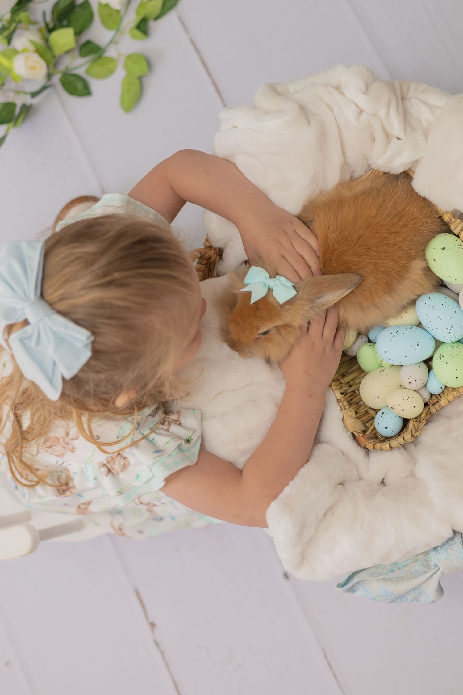 A person pets a ginger cat while sitting on a white blanket next to pastel Easter eggs in a spring setting.