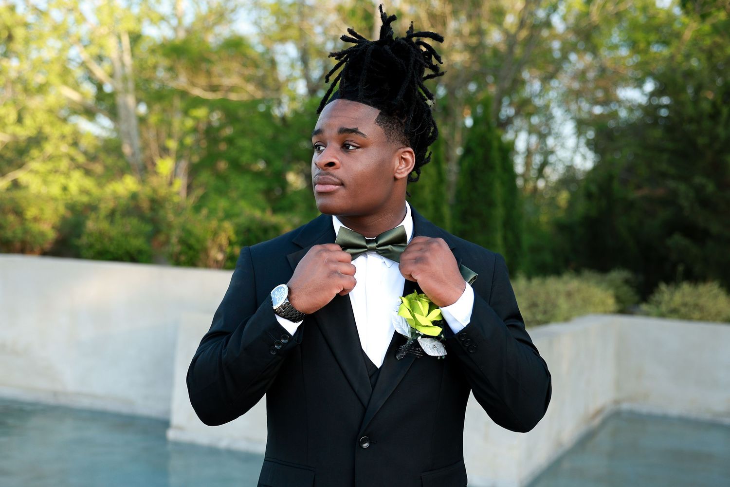 Young man in black tuxedo adjusting bow tie outdoors, wearing boutonniere, dressed for prom or formal event.