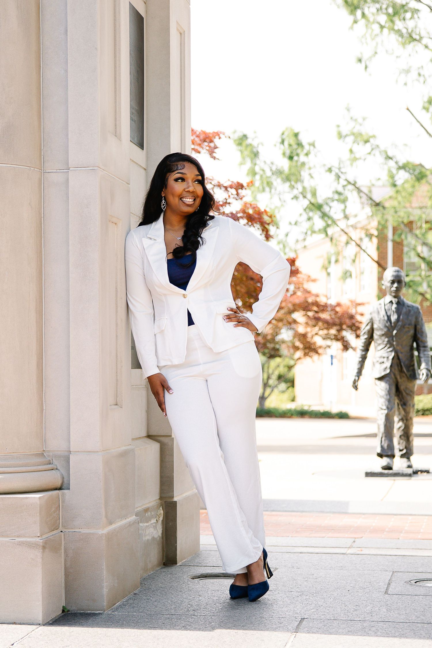 Woman in white pantsuit posing confidently against a stone column outdoors near a bronze statue.