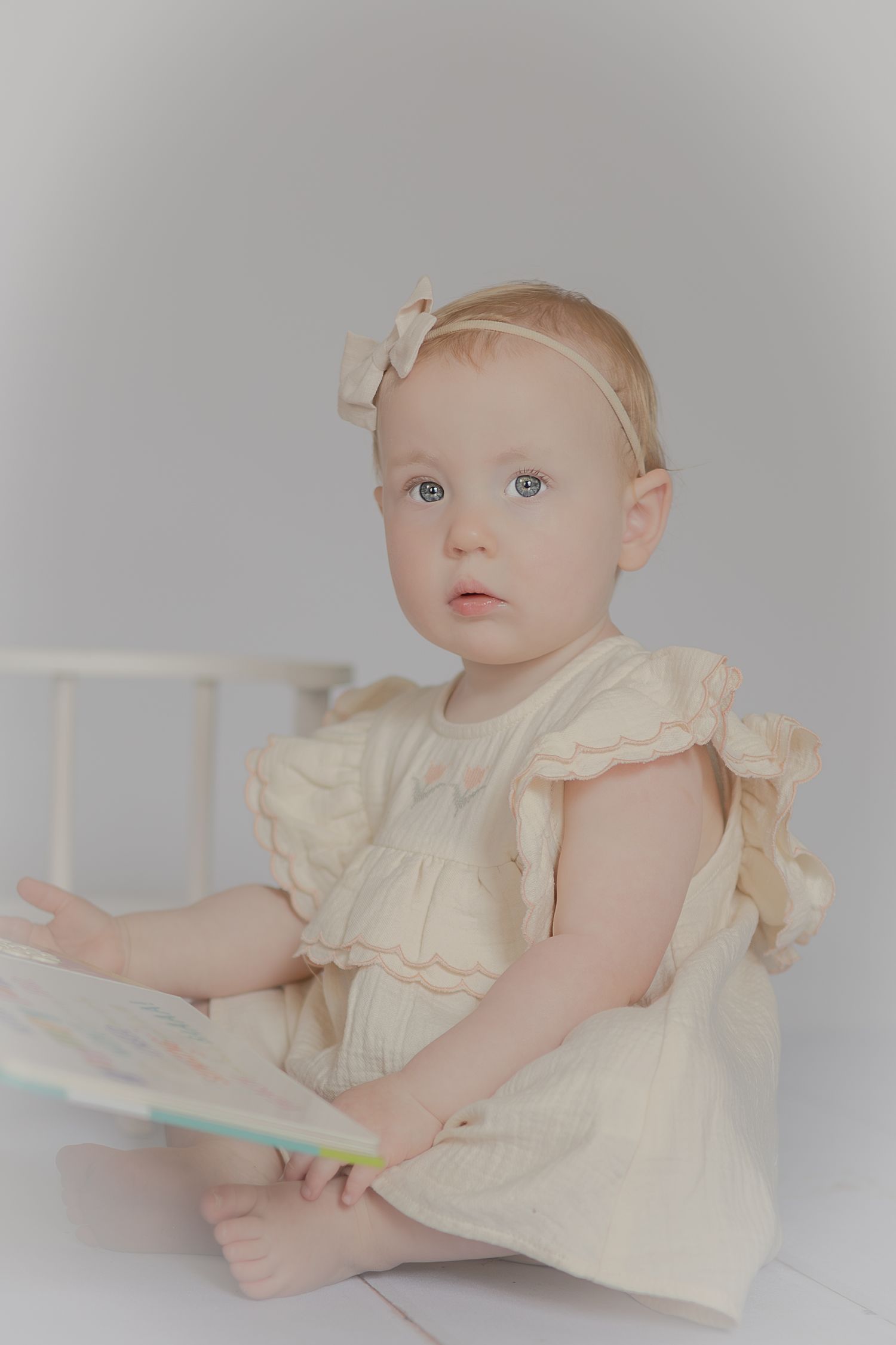 Adorable baby girl in cream ruffle dress and bow headband sitting on white floor holding a colorful book.