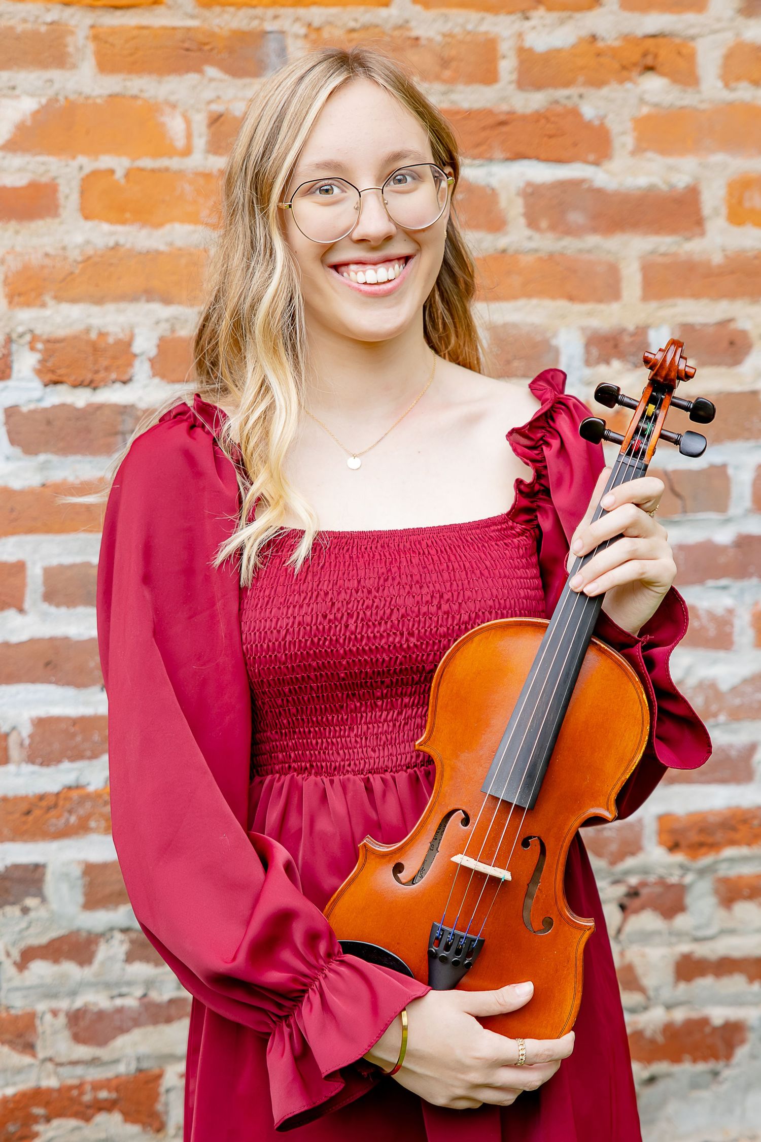 Smiling blonde woman in red dress holding a violin against a brick wall background.