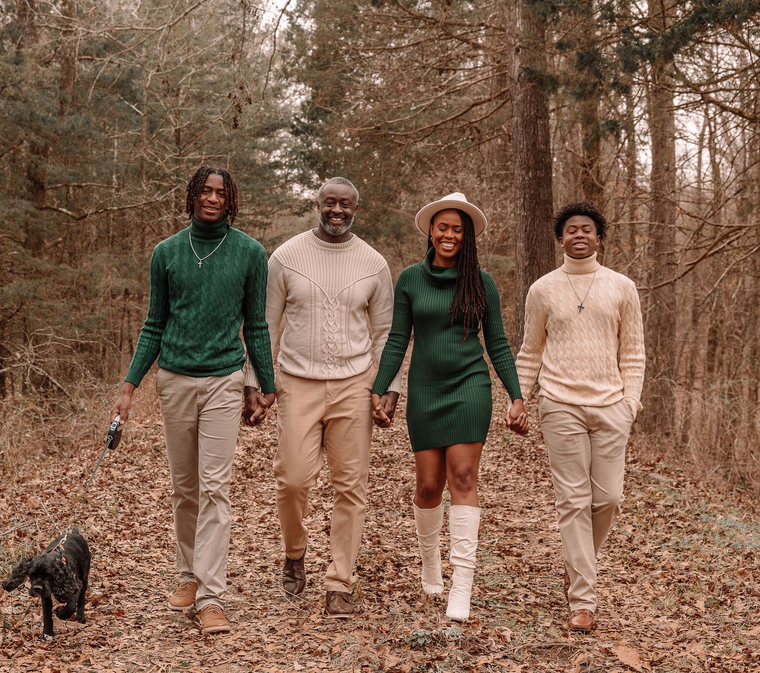 Four people in coordinated green and beige outfits walking together with a small black dog on a wooded autumn trail.