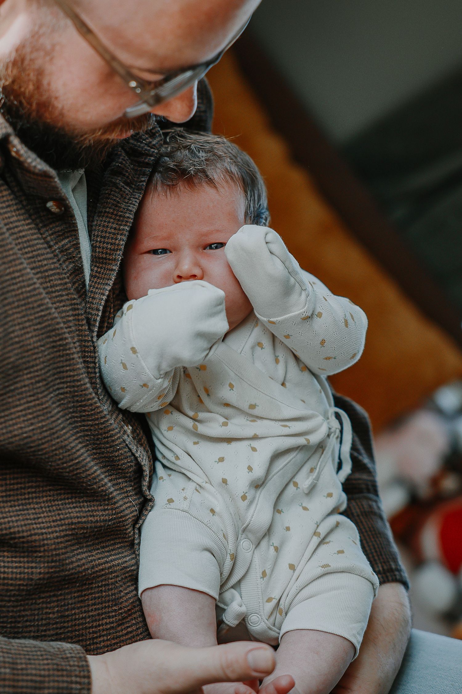 A parent in a brown sweater holds a baby wearing a light-colored onesie with small patterns during a tender moment.