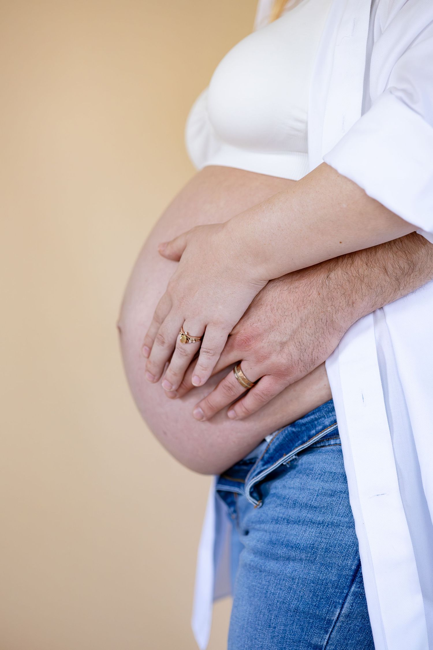 Close-up view of hands with wedding rings resting on a pregnant belly, wearing white shirt and jeans.