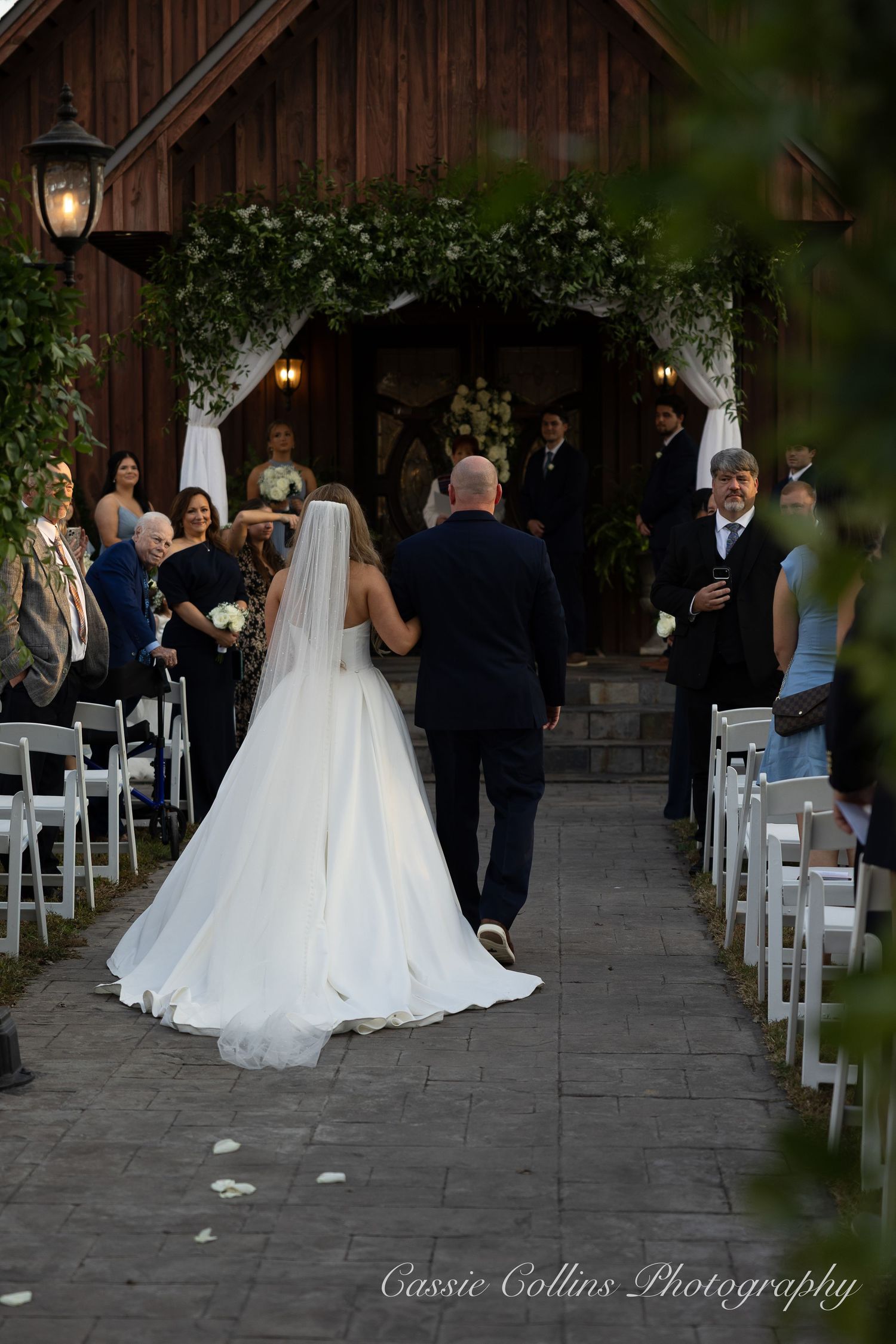 A bride in a white gown walks down an outdoor aisle lined with white chairs at a rustic barn wedding ceremony.