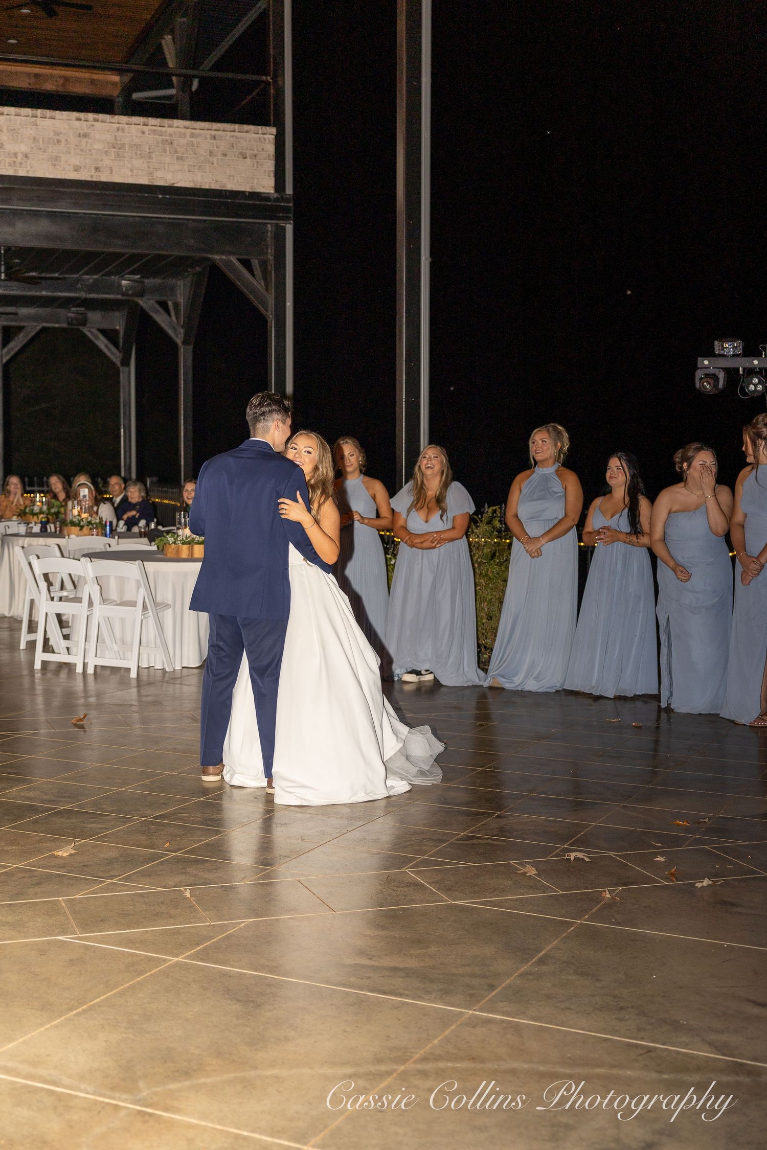 A couple shares their first dance at a wedding reception while bridesmaids in light blue dresses watch from the side.