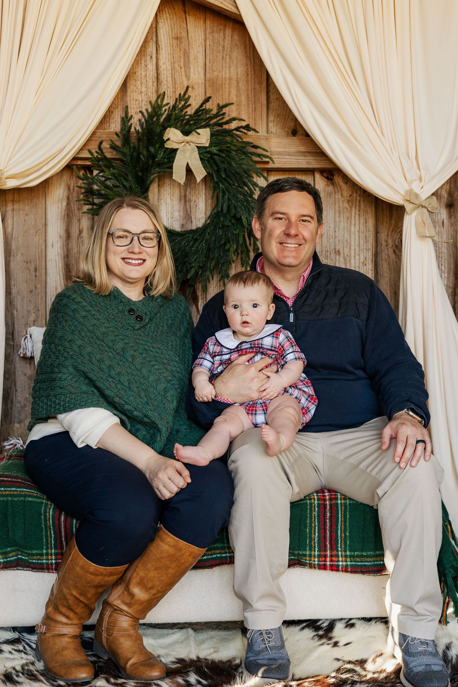 Family holiday portrait on rustic bench with evergreen wreath and cream curtains against wooden backdrop.
