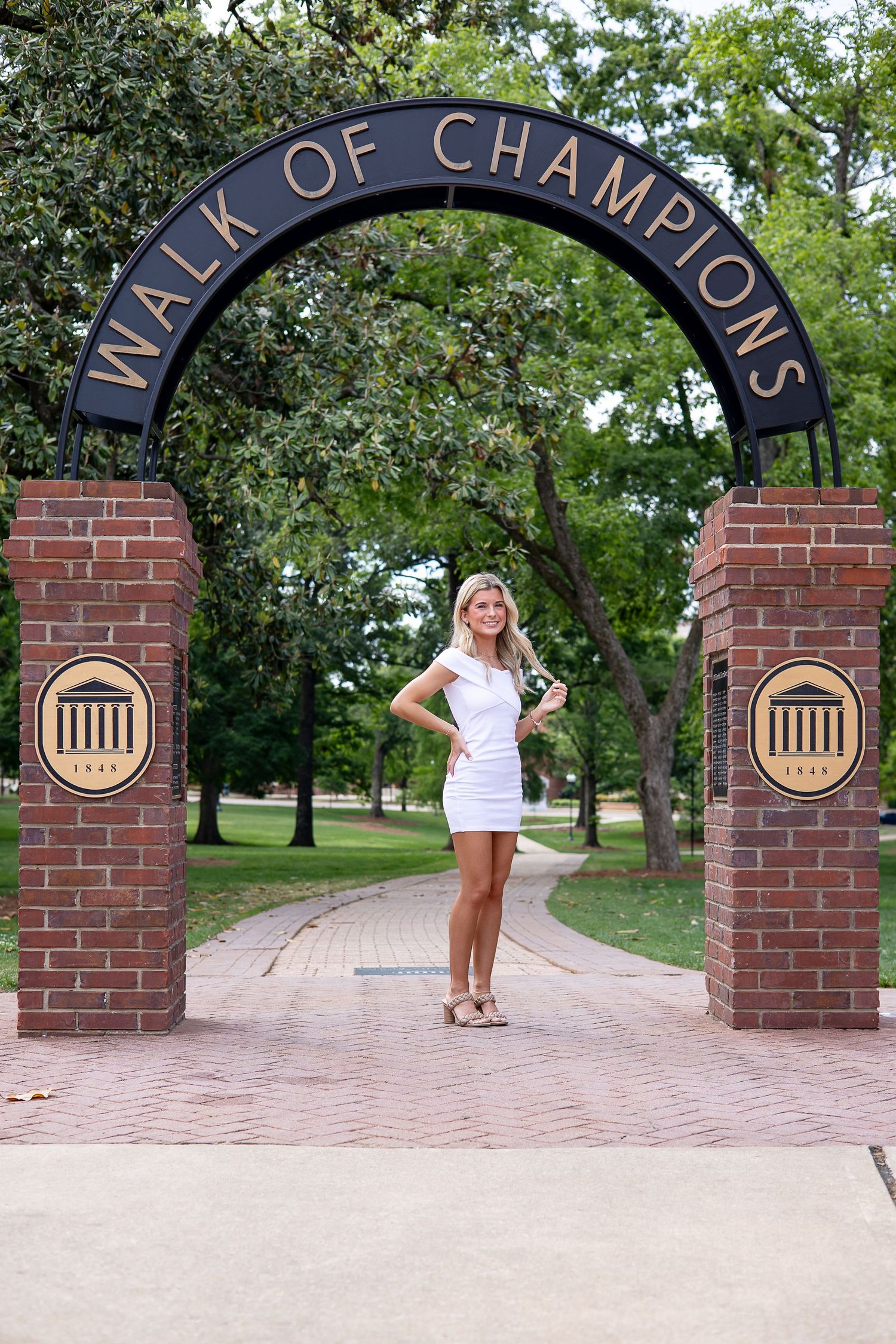 Smiling blonde woman in white dress posing under the Walk of Champions arch at Ole Miss university.