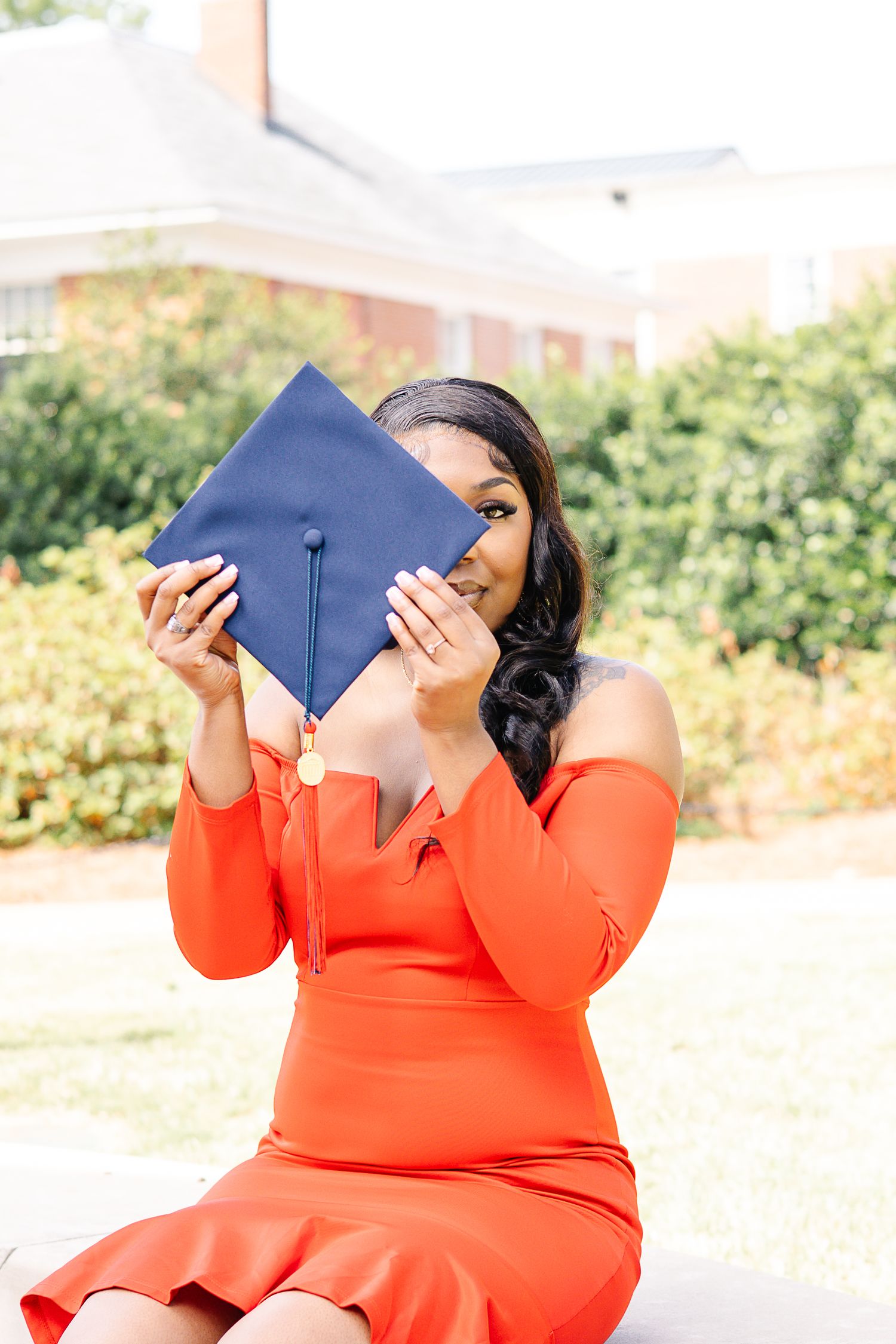 A young woman in an orange off-shoulder dress holds a navy blue graduation cap in front of her face outdoors.