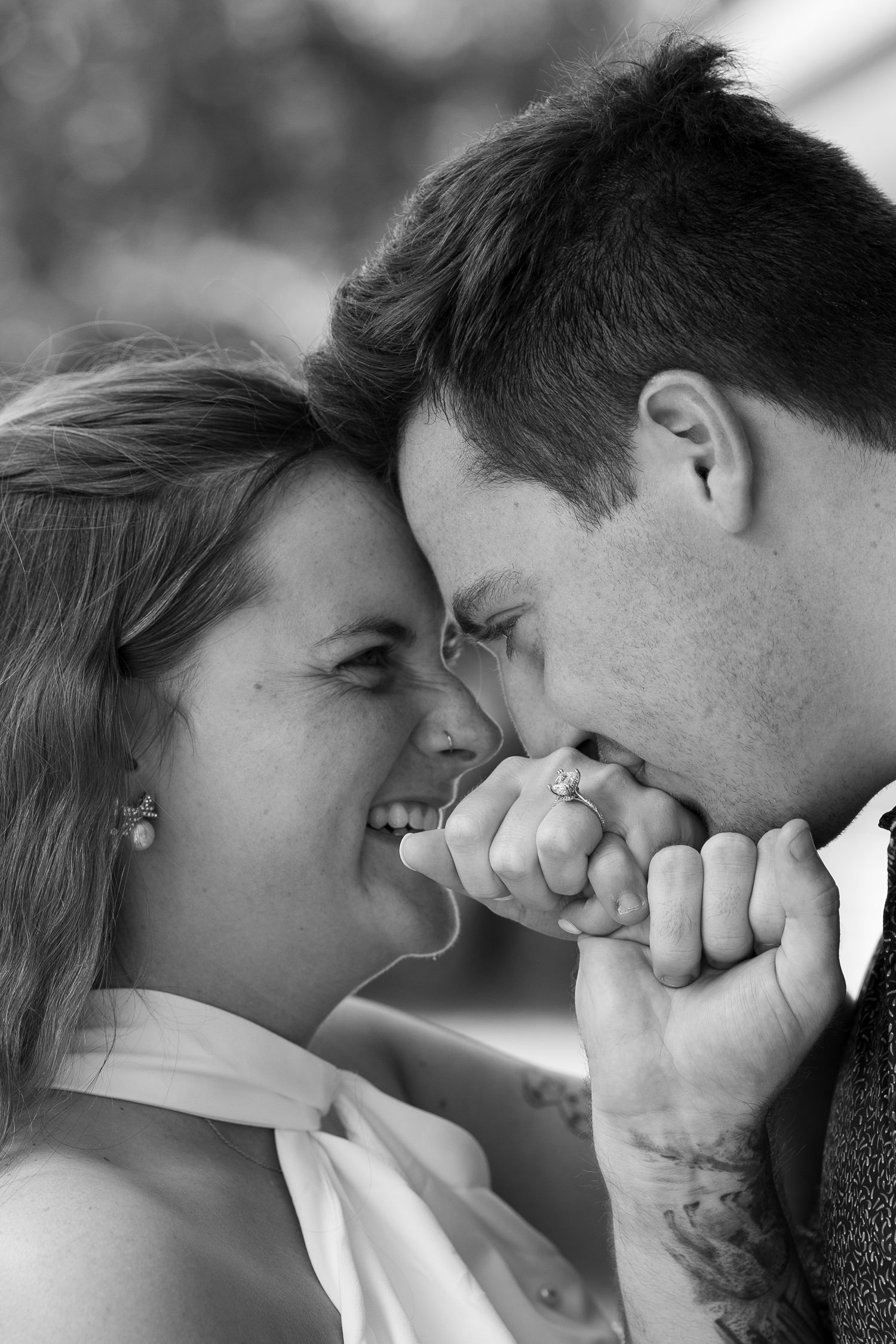 Black and white photo of a smiling couple touching foreheads, man kissing womans hand with engagement ring.