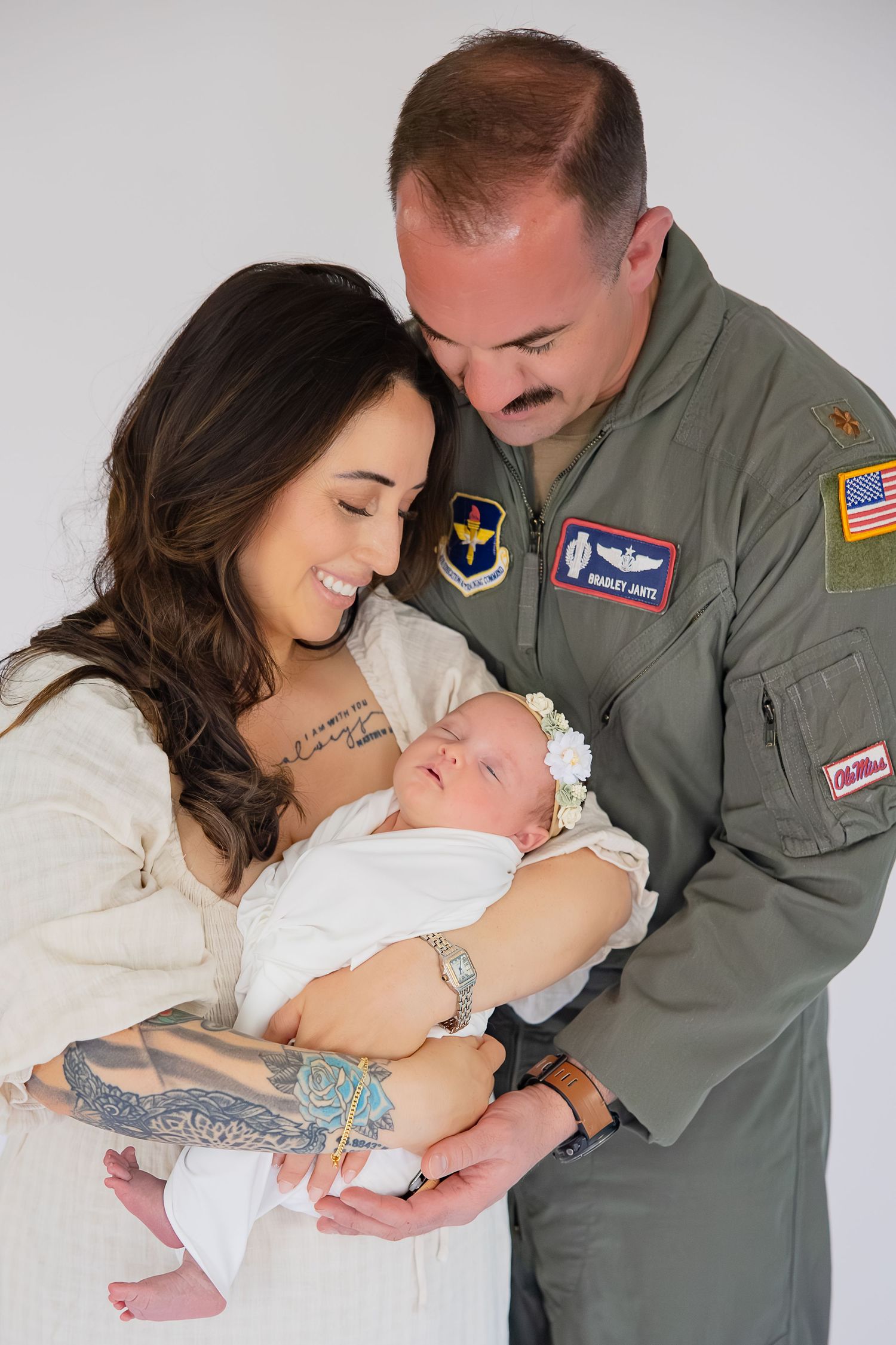 Military pilot in flight suit holds newborn baby with smiling brunette woman in white dress in studio portrait.