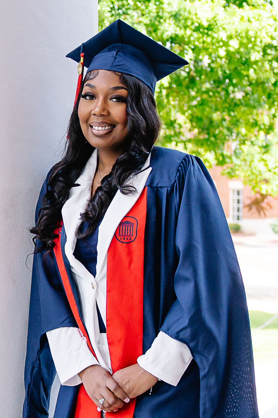 Smiling graduate in navy cap and gown with red and white stole poses outdoors near a white column on campus.