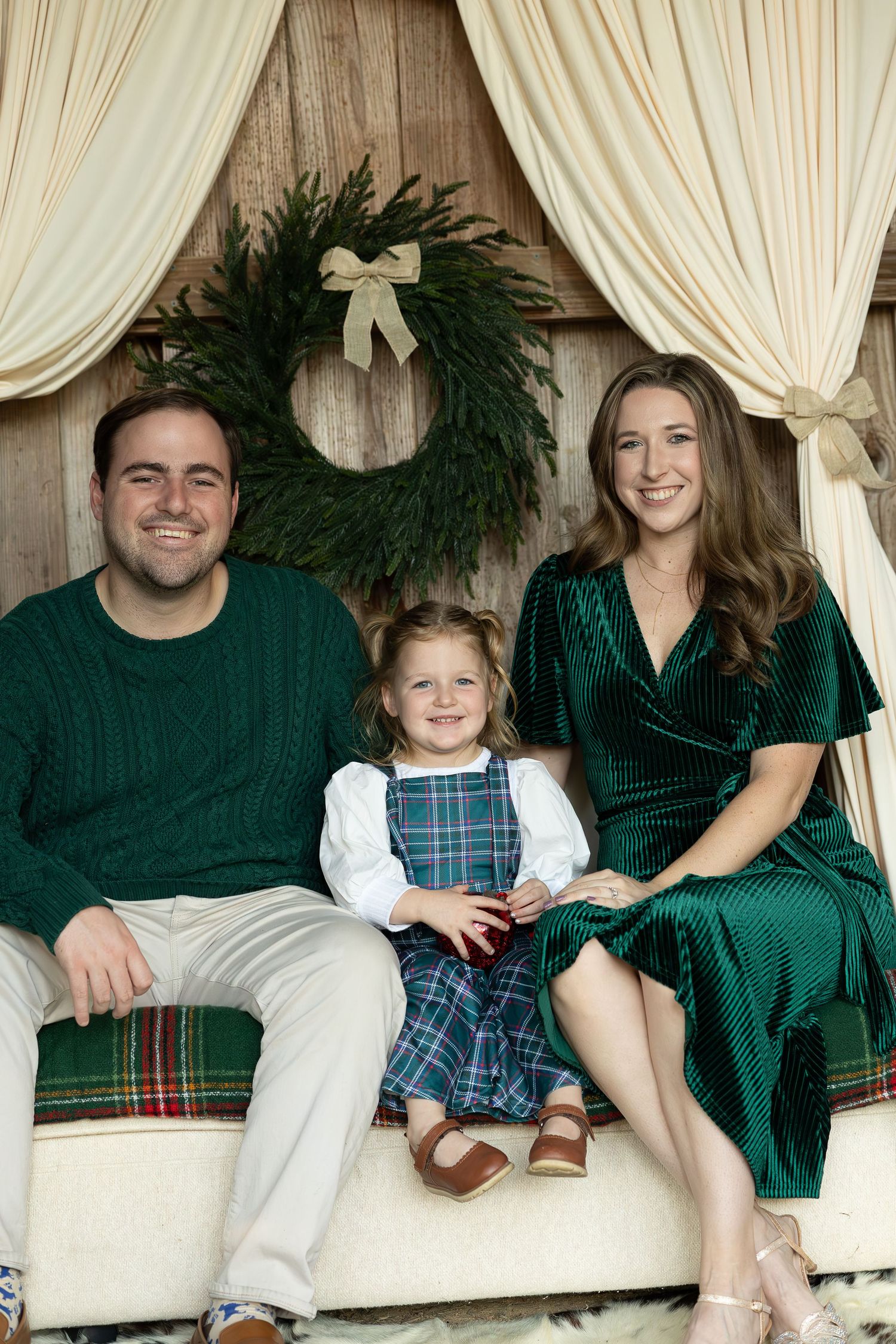 A family in coordinated green and plaid holiday outfits sits together on a cream-colored couch with draped fabric backdrop.