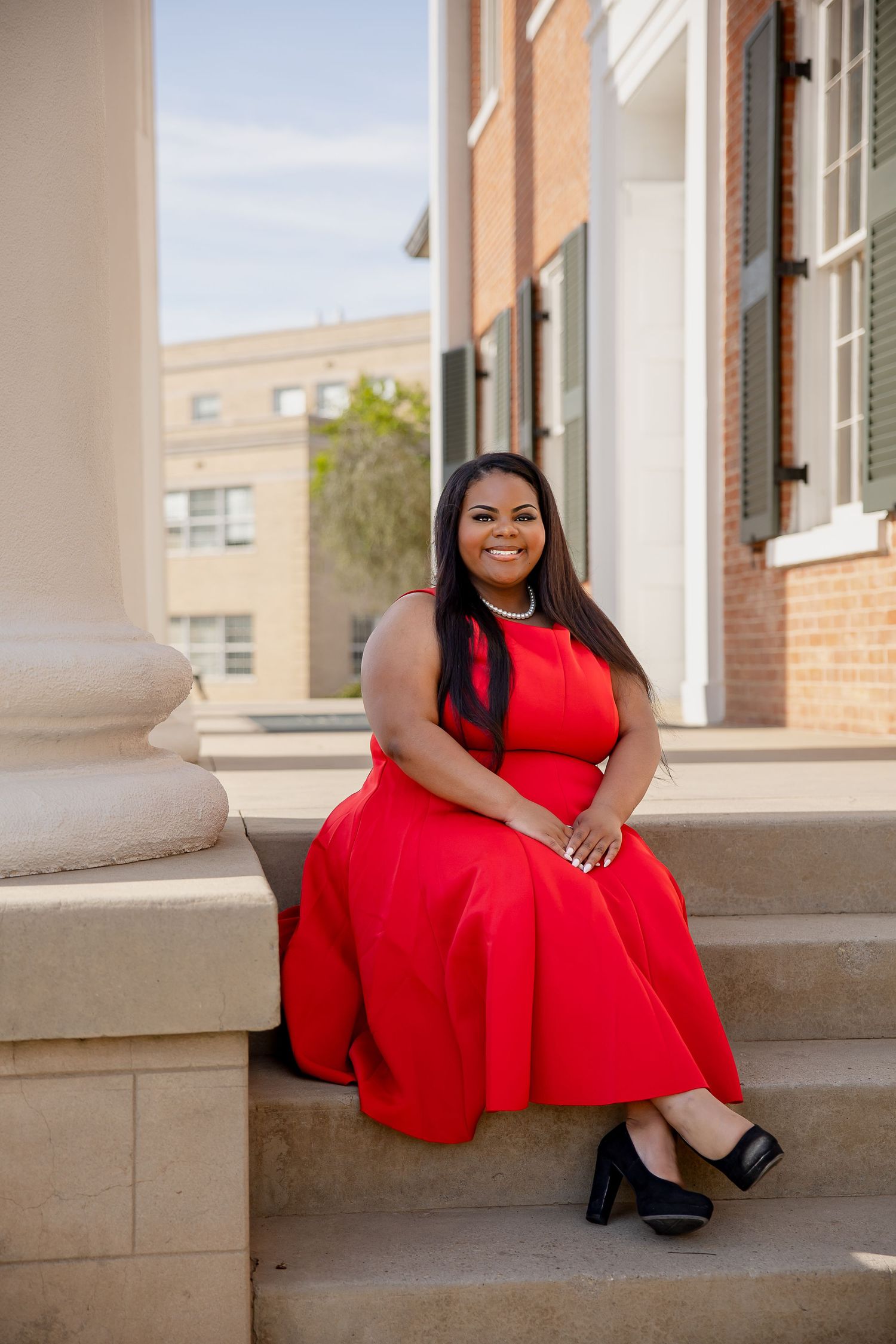 Woman in red dress sitting on stone steps outside brick building, smiling confidently in elegant sleeveless gown and black heels.