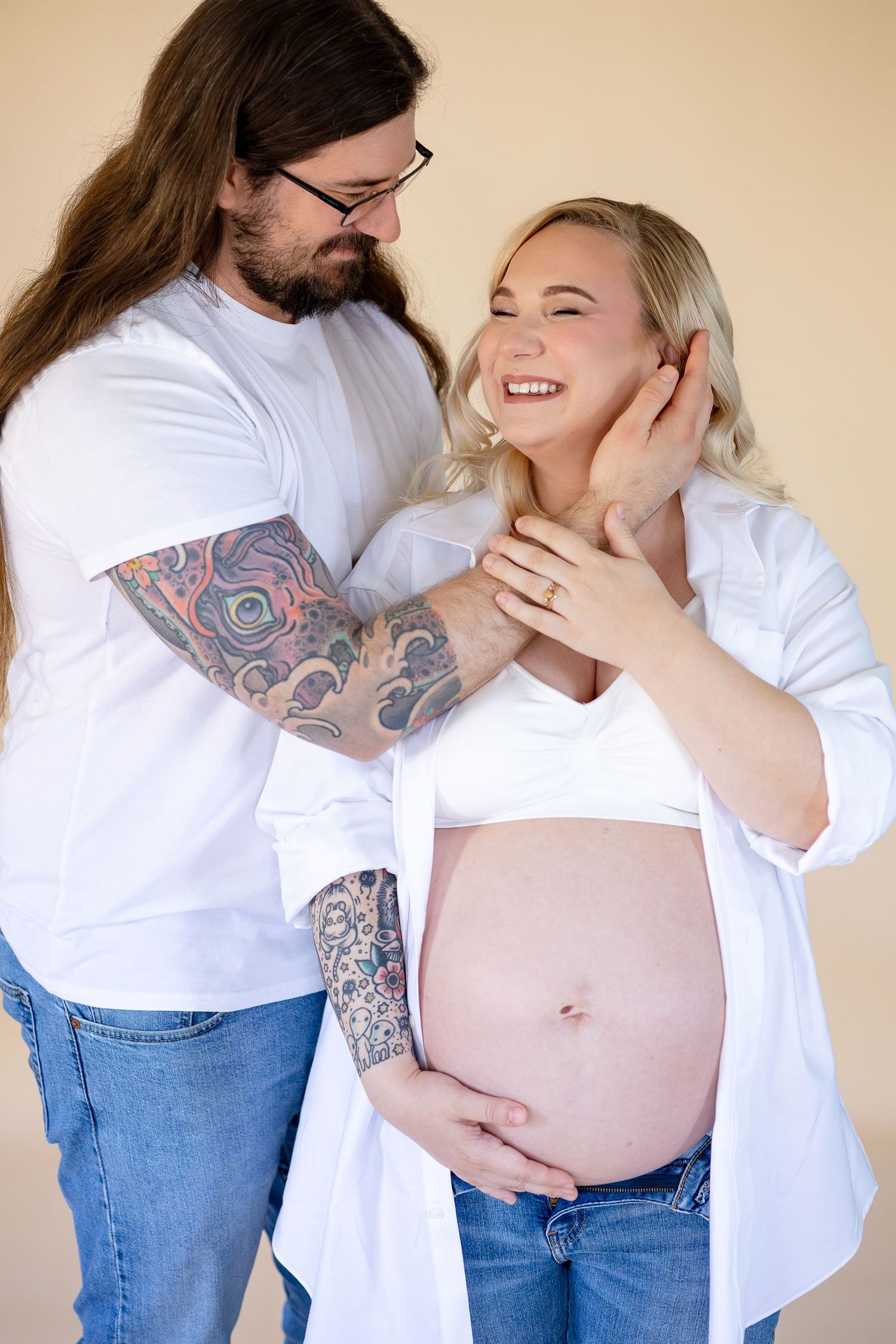 A couple embraces during a pregnancy photoshoot, wearing white shirts and jeans against a neutral backdrop.