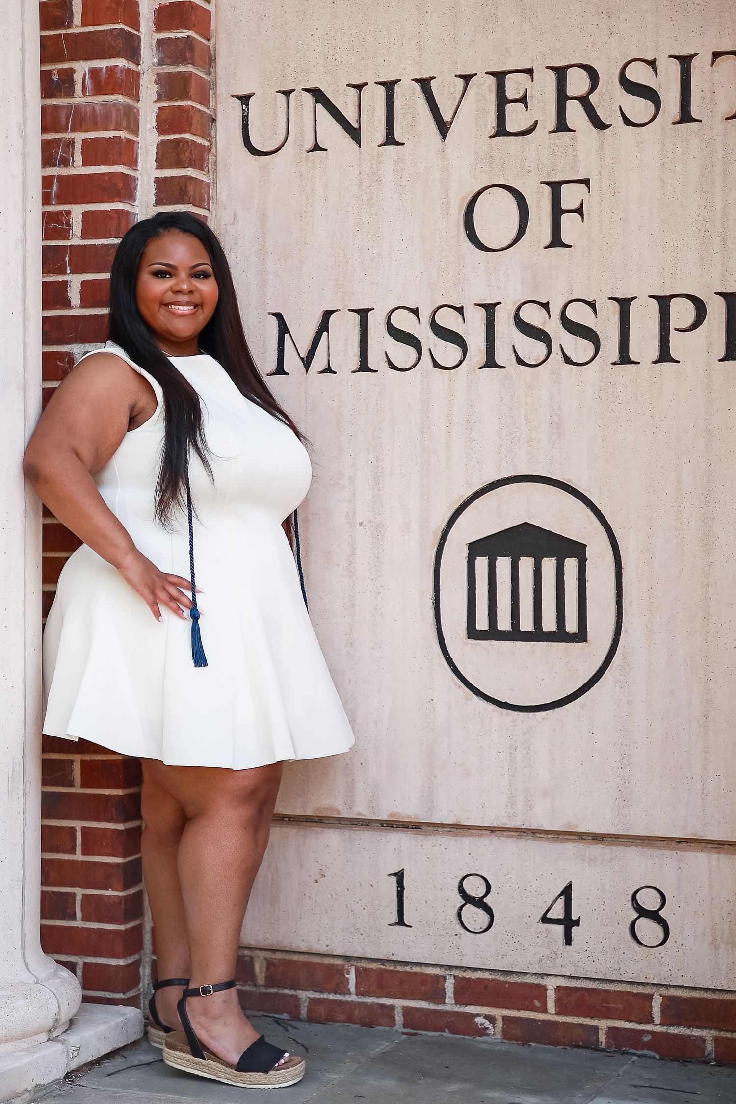 Young woman in white dress posing by University of Mississippi 1848 sign during graduation celebration.
