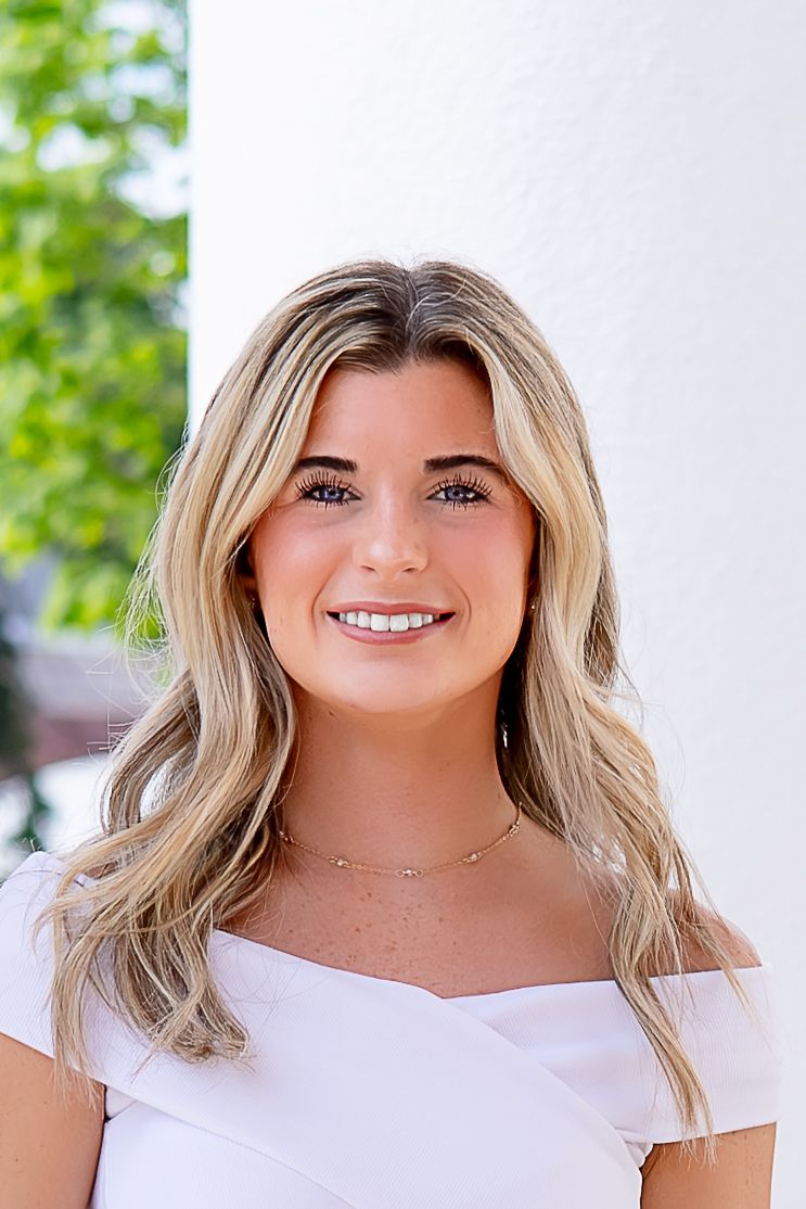 Young blonde woman smiling in white off-shoulder top, posing outdoors near white column with green foliage background.