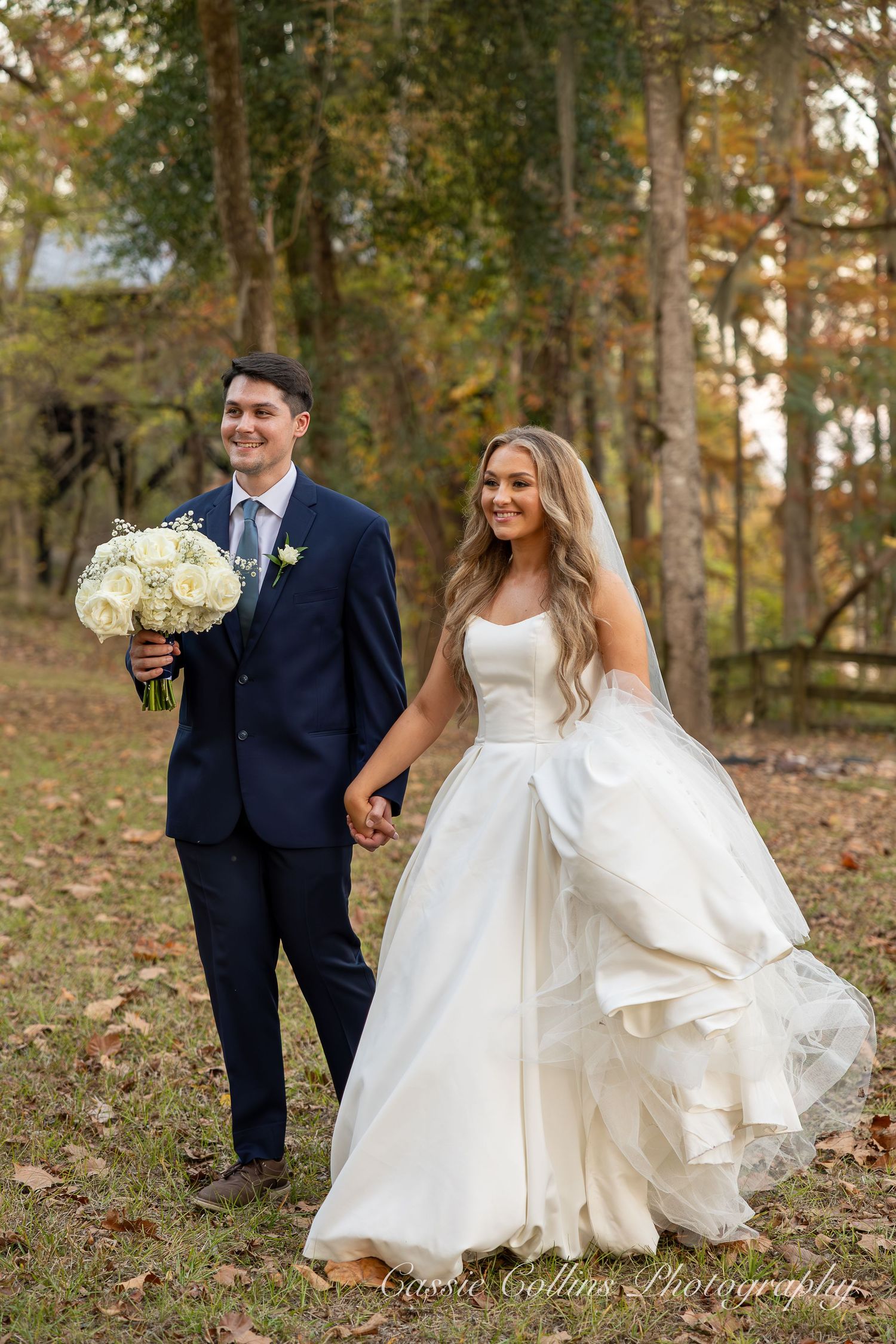 A couple walks together outdoors on their wedding day, with the bride's white dress flowing in the autumn breeze.