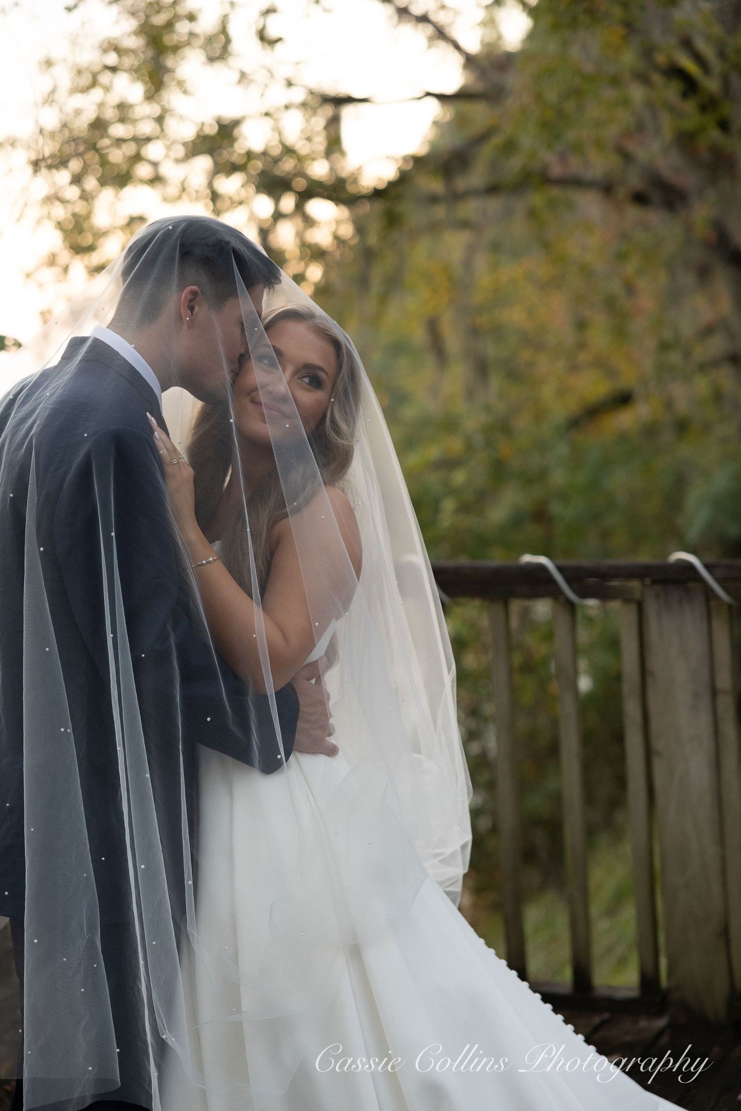 Bride and groom share romantic moment under wedding veil on wooden bridge surrounded by autumn foliage.