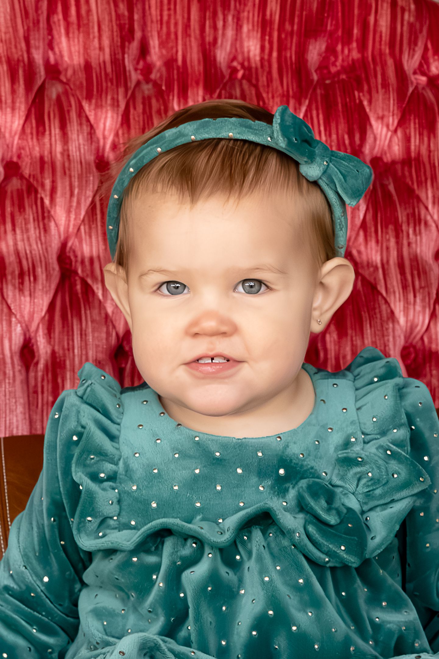 A baby wearing a teal blue polka dot dress with ruffles and matching headband poses against a red textured backdrop.