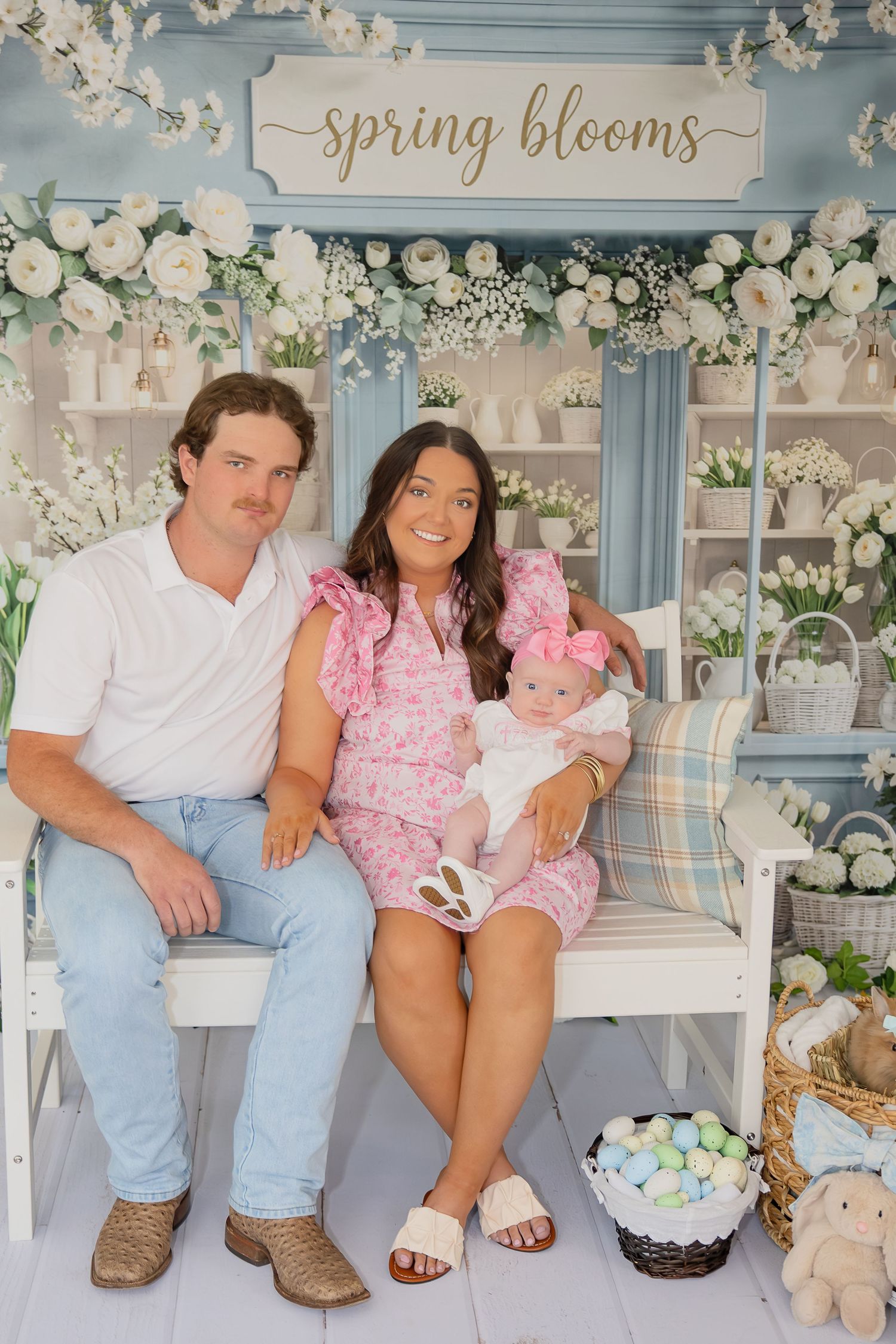 Family spring photo session with parents and baby on white bench, floral backdrop with spring blooms sign.