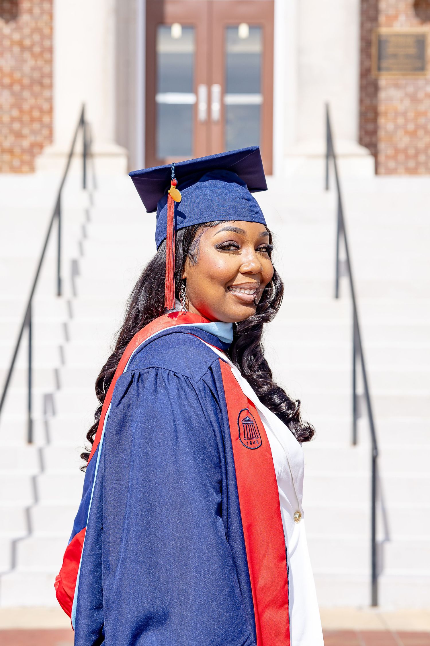 Smiling graduate in blue cap and gown with red sash poses on university steps on graduation day.