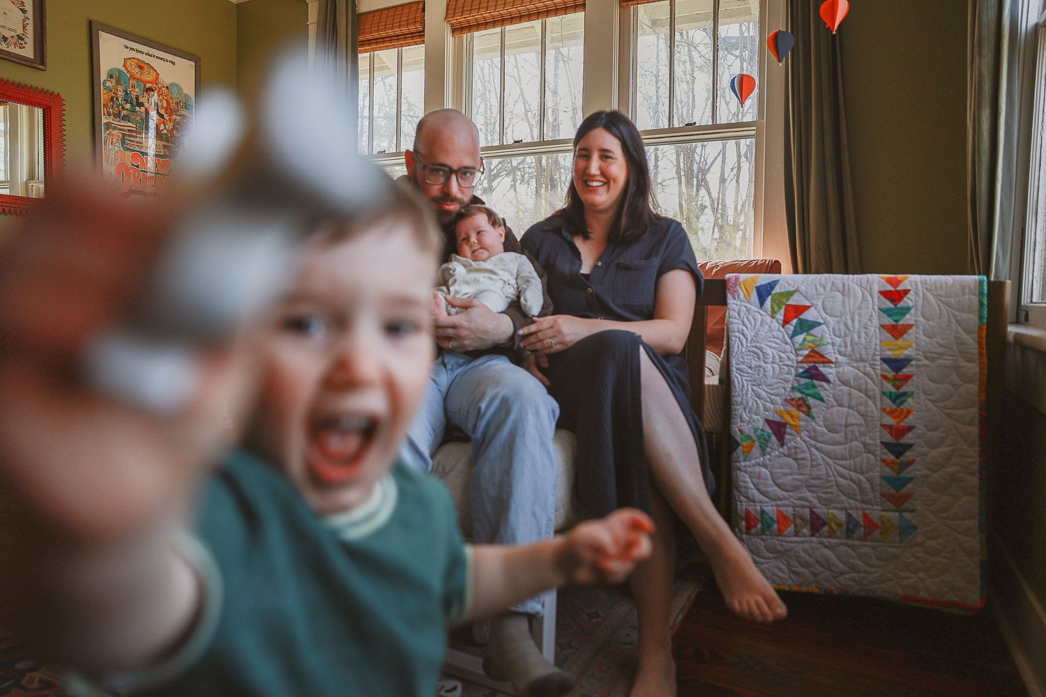 Happy family with two young children laughing and playing together in a cozy living room with a colorful quilt.