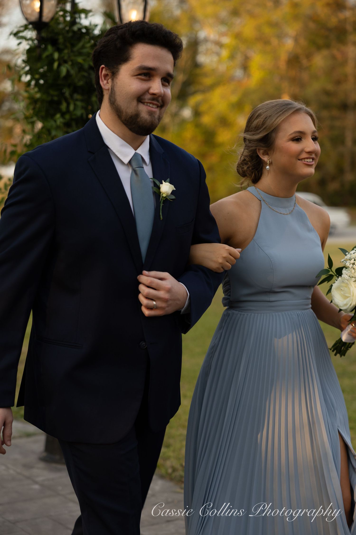 Wedding couple walks arm in arm outdoors with the bridesmaid wearing a flowing dusty blue halter dress.