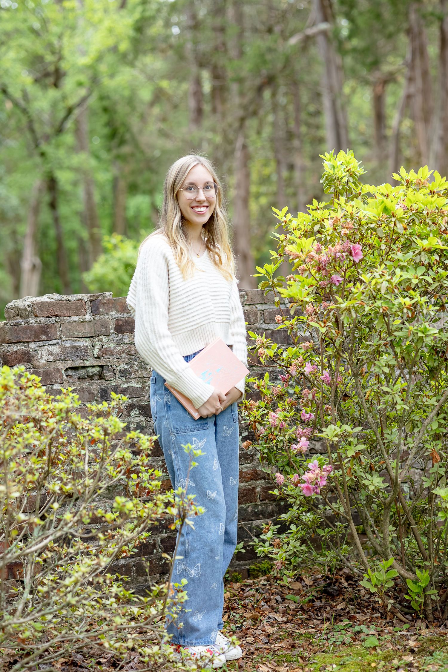 Smiling young blonde woman in cream sweater and jeans holding a book beside pink azalea blooms in a garden.