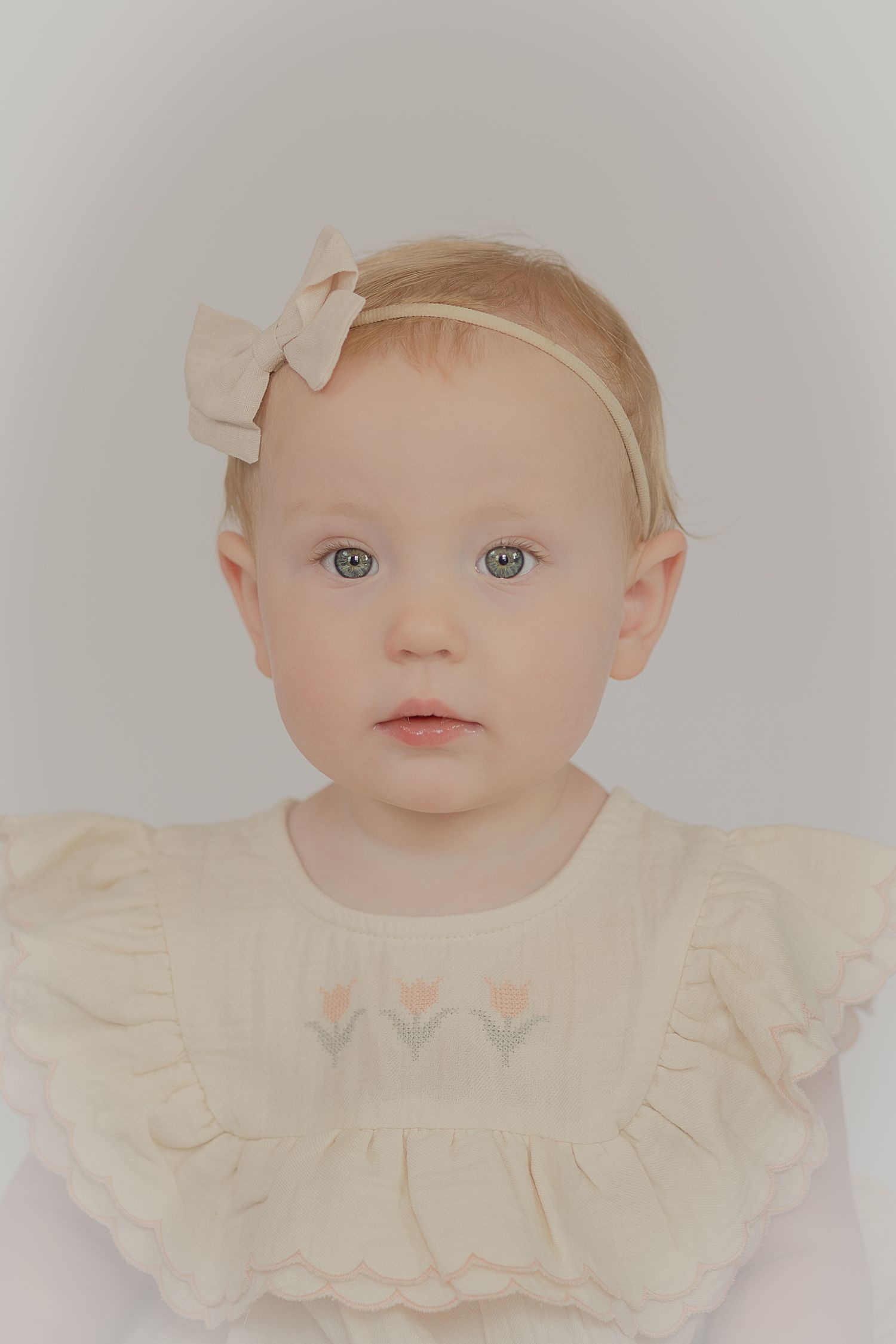 Baby girl with blue eyes wearing a cream dress and bow headband poses for a soft, vintage-style portrait photo.