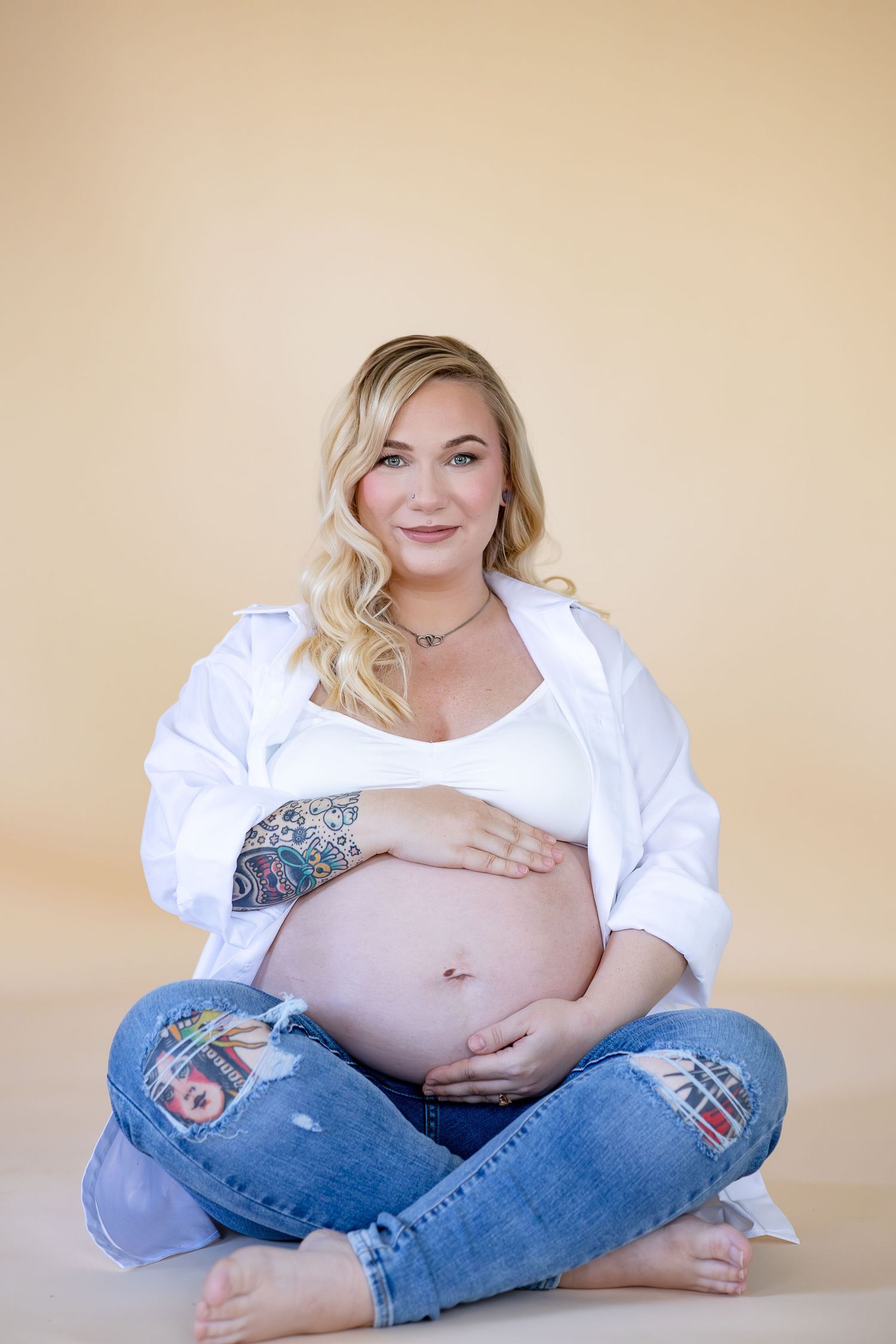 A maternity photo series showing a woman in white shirt and jeans sitting cross-legged against beige background.