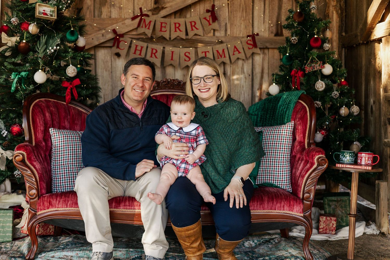 Family poses for holiday photos in a rustic barn setting with Christmas decorations and a festive tree.