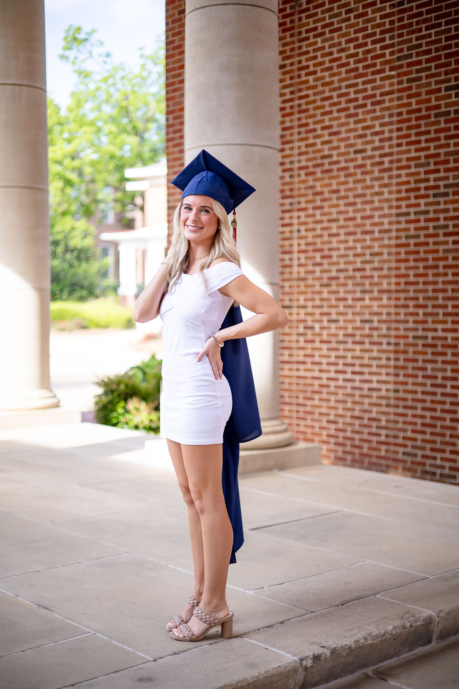 Smiling blonde graduate in white dress and navy cap and gown posing outdoors near brick columns.