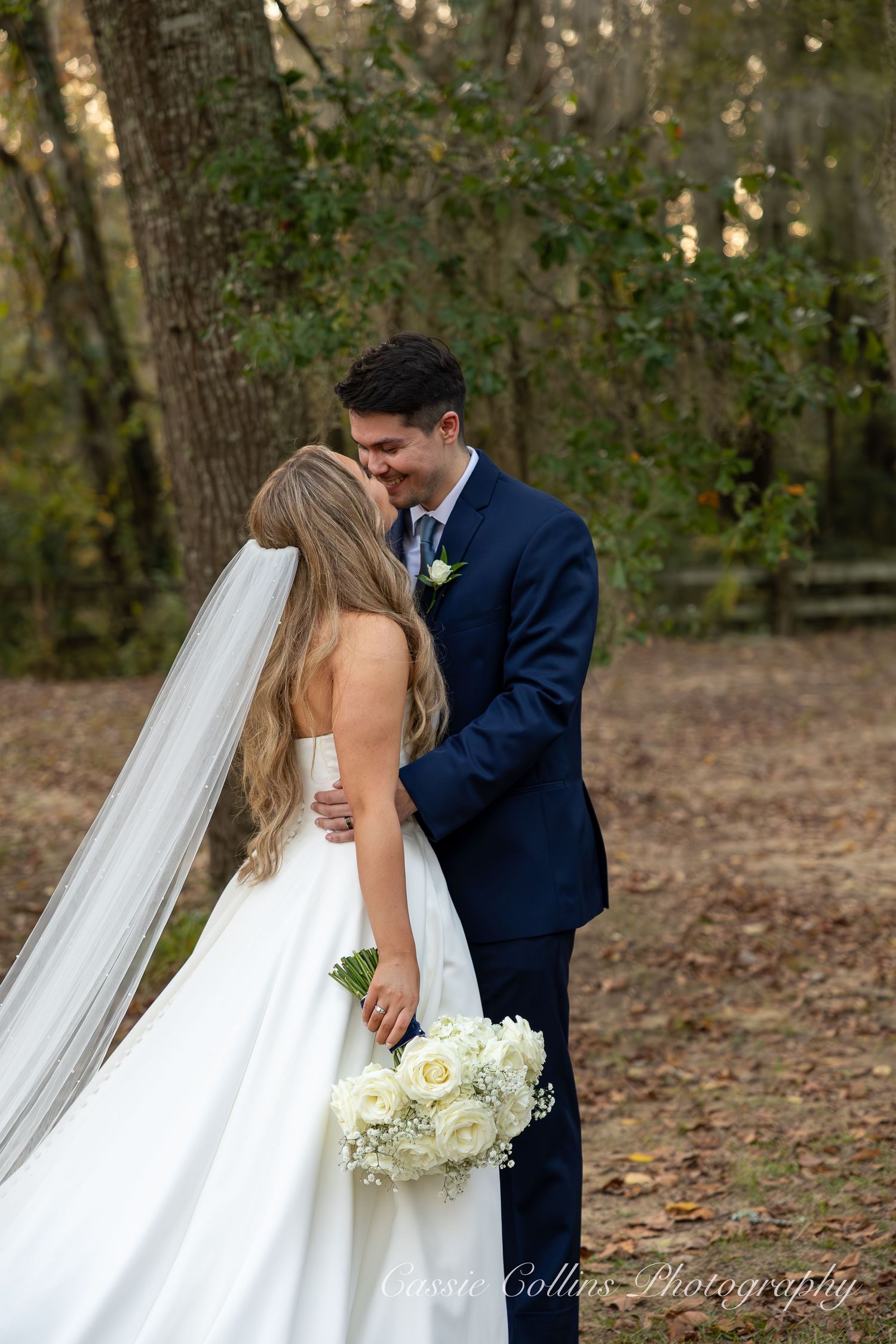 A romantic outdoor wedding moment captured in a wooded setting with a flowing white gown and navy suit.